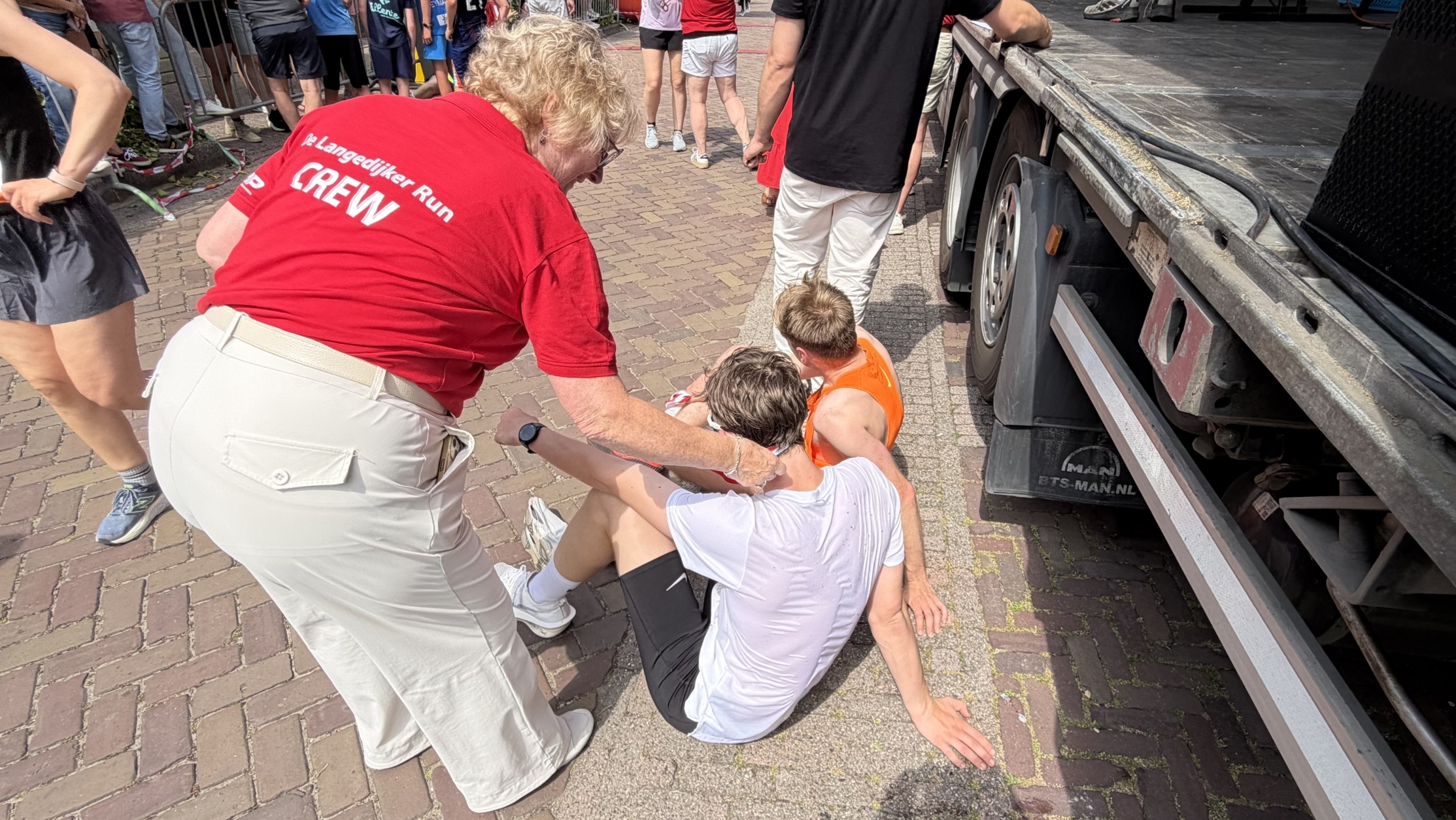 Een vrouw in een rood T-shirt met "CREW" erop helpt twee zittende deelnemers bij een evenement naast een vrachtwagen.