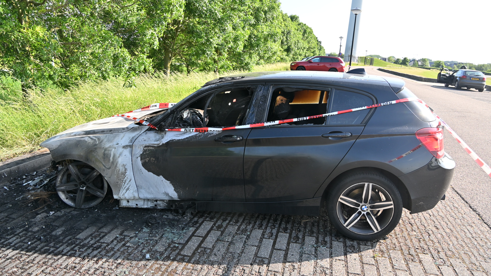 Zwarte auto met brandschade aan de voorzijde, omringd door rood-wit politietape naast een grasveld.