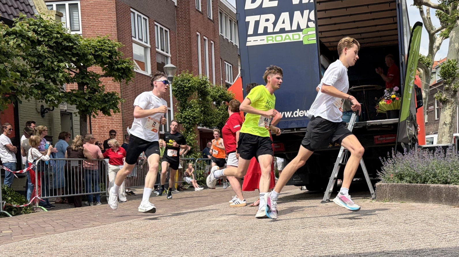 Marathonlopers rennen langs een straat waar toeschouwers langs de kant staan, terwijl een vrouw op een verhoogd gedeelte zit.