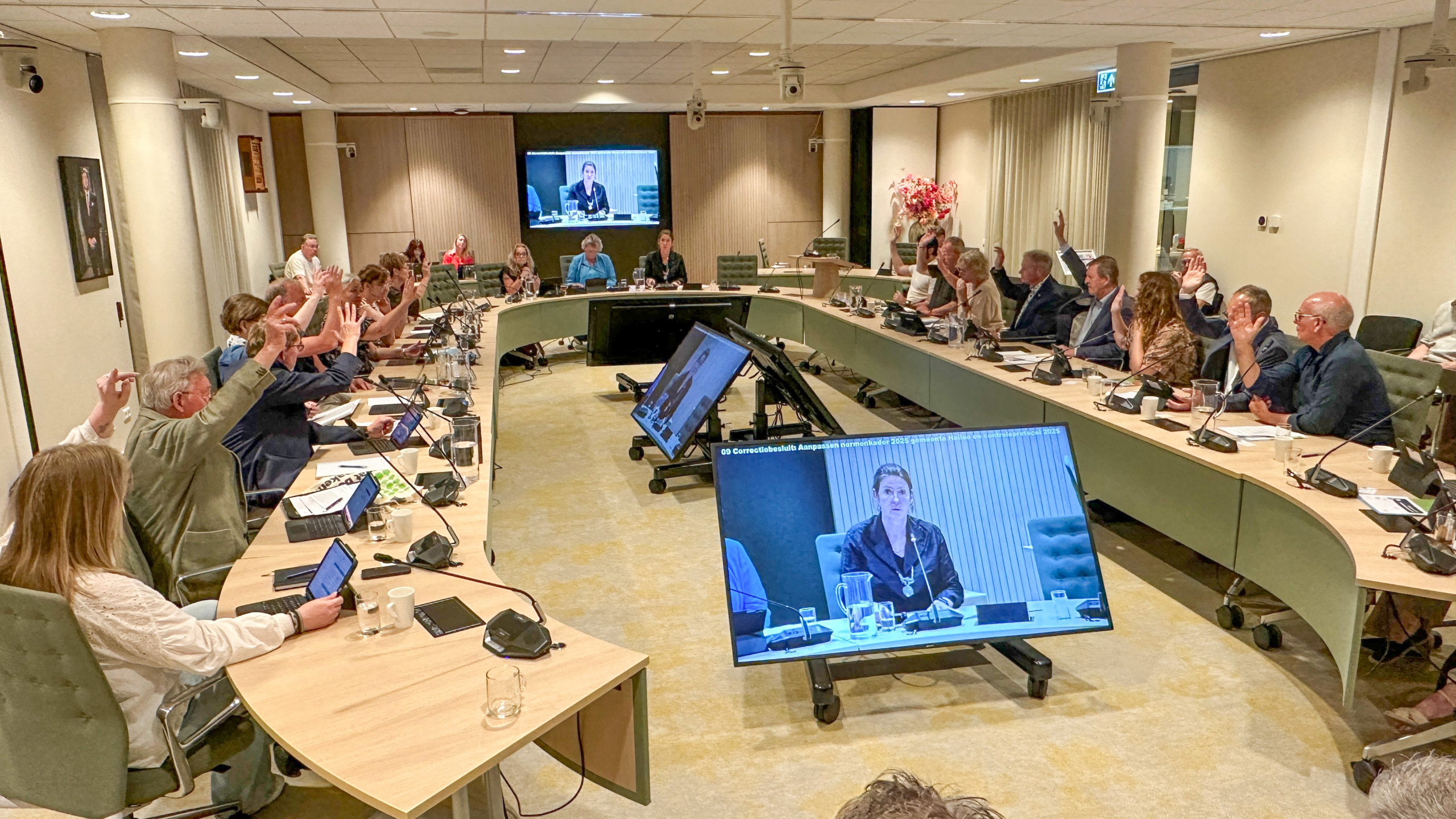 Mensen in een vergaderzaal stemmen met opgestoken handen; een spreker verschijnt op een scherm.
