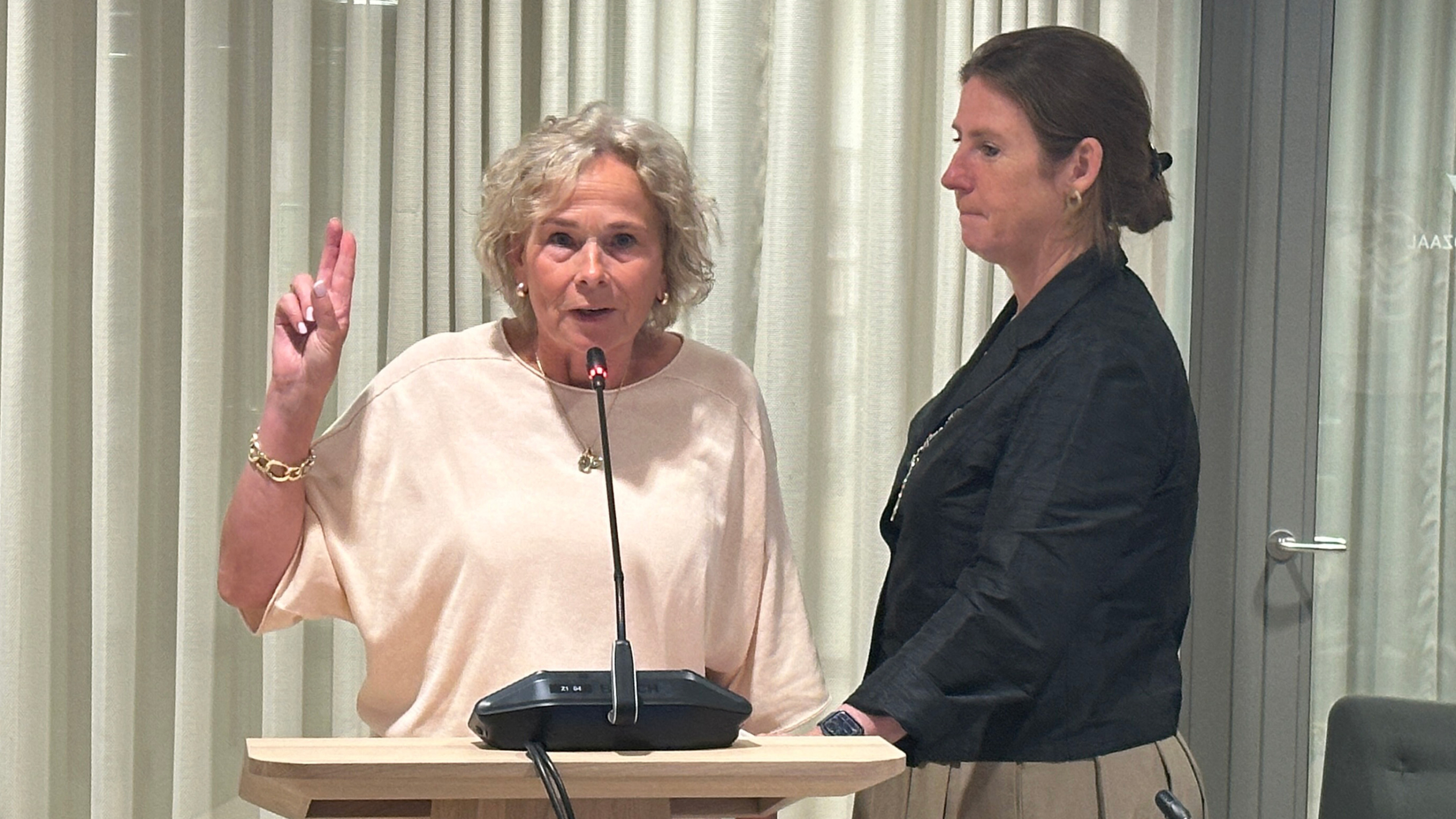 Twee vrouwen staan bij een microfoon, één met opgeheven hand, in een officiële setting met gordijnen op de achtergrond.