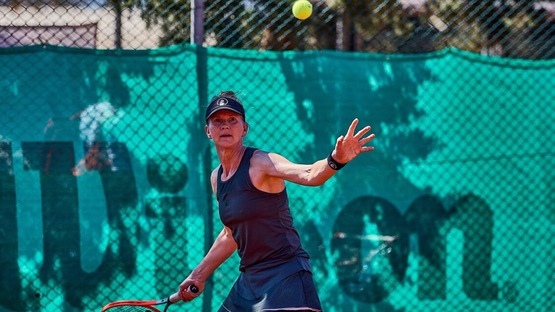 Een vrouw speelt tennis op een buitenbaan, klaar om de bal met haar racket te slaan tegen een groene achtergrond.