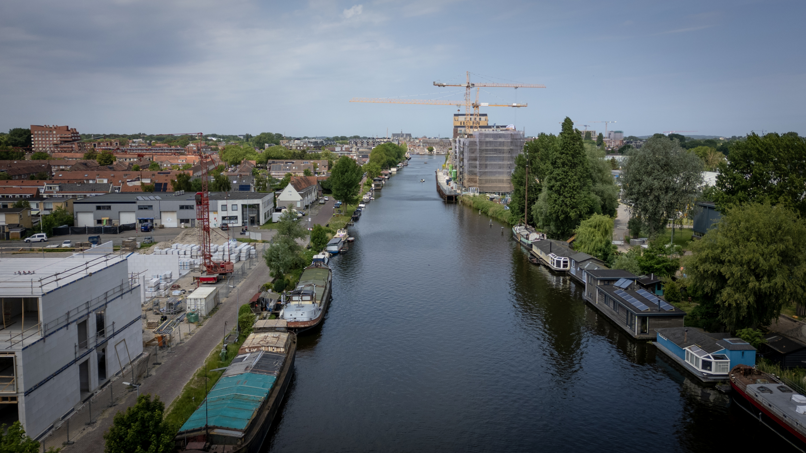 Kanaal met woonboten en een bouwterrein met een kraan aan de linkerkant, omgeven door groene bomen en huizen.