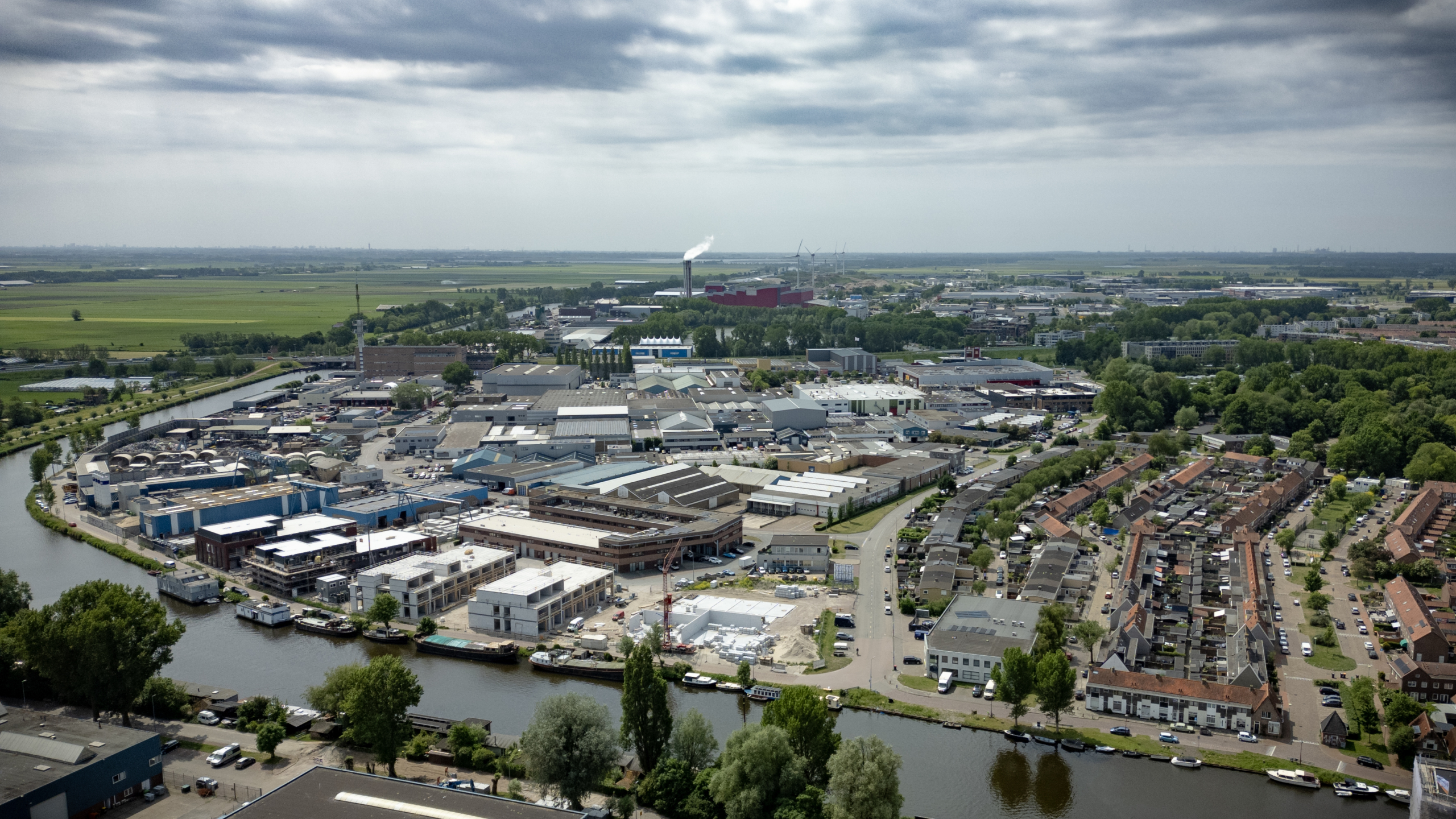 Luchtfoto van een industriegebied met fabrieken en een woonwijk ernaast, omgeven door groen en sloten onder een bewolkte hemel.