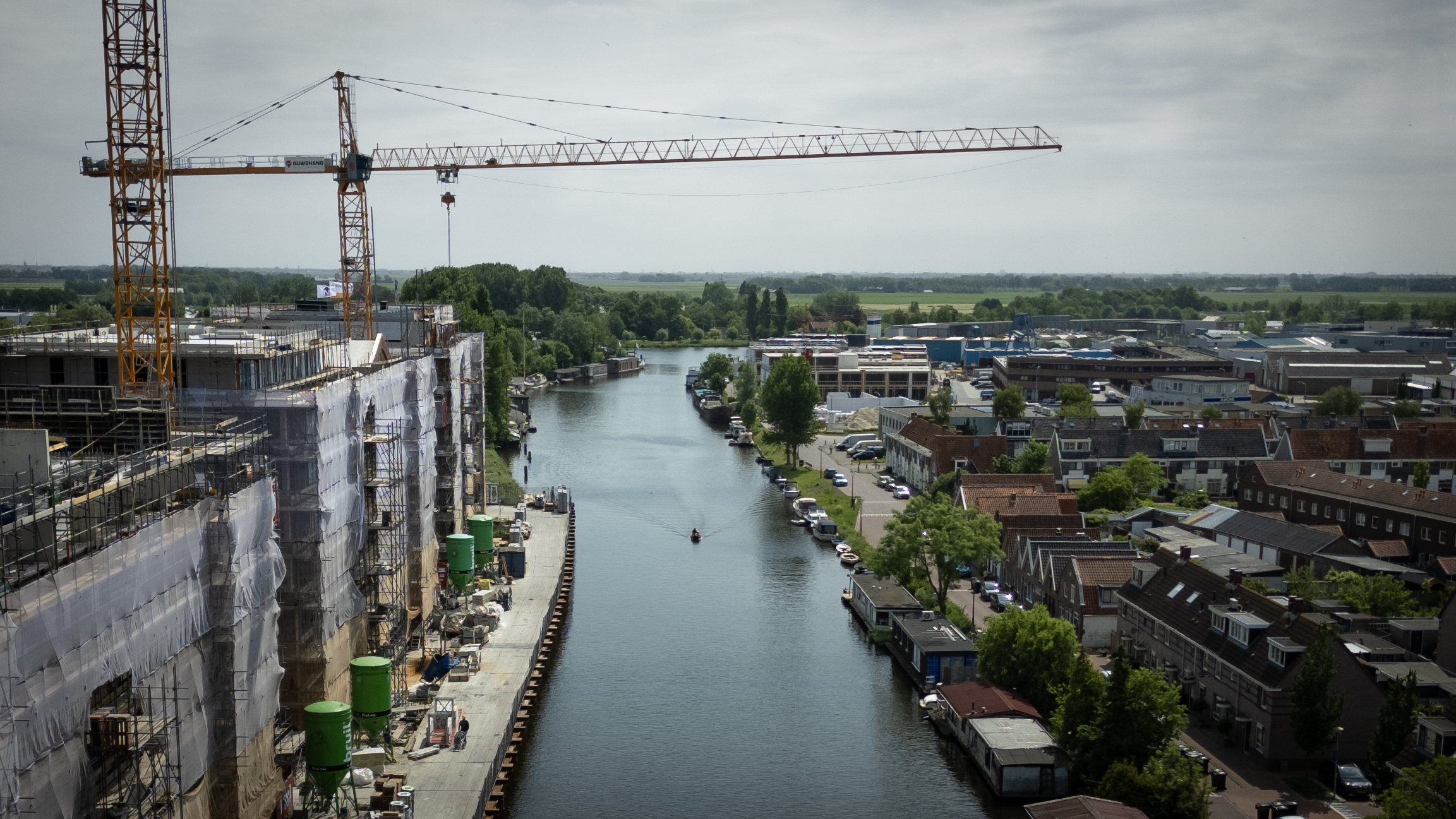 Een bouwplaats aan een kanaal met hijskranen en steigers; woningen en groene bomen langs het water.