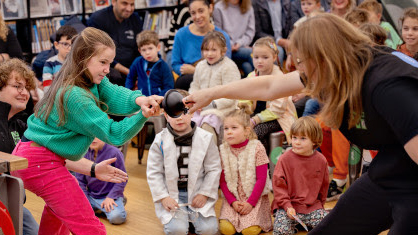 Een kind in een groene trui neemt enthousiast deel aan een interactieve activiteit met een volwassene, terwijl andere kinderen toekijken.