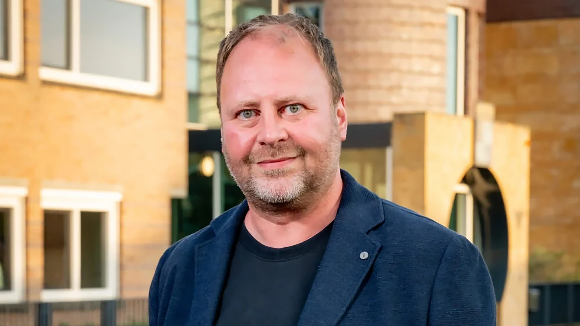 Man in een donkerblauwe blazer staat buiten voor een gebouw met bakstenen muren en ramen.