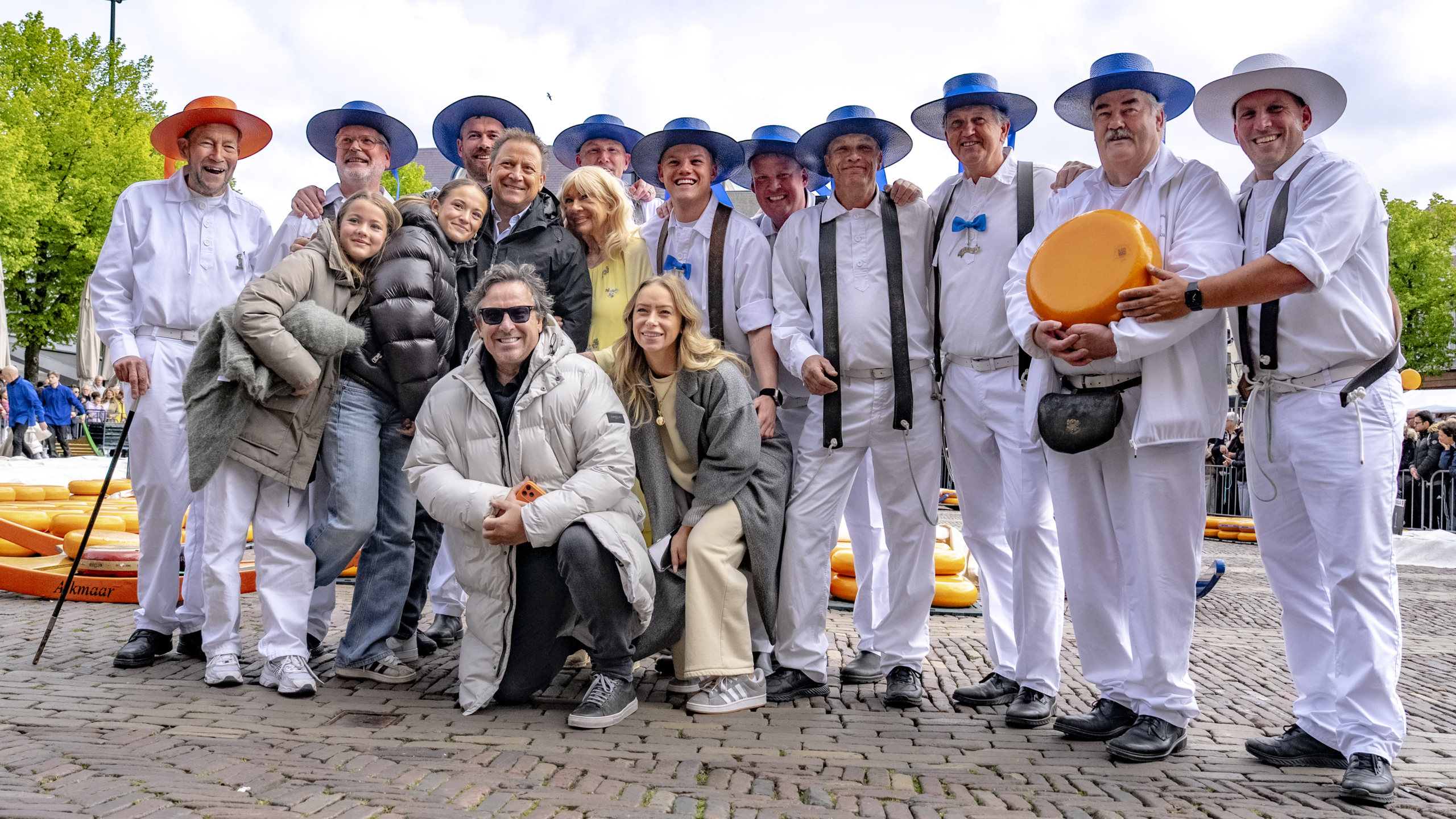 Een groep mensen poseert samen, sommigen in traditionele witte kleding en gekleurde hoeden, naast grote ronde kazen.