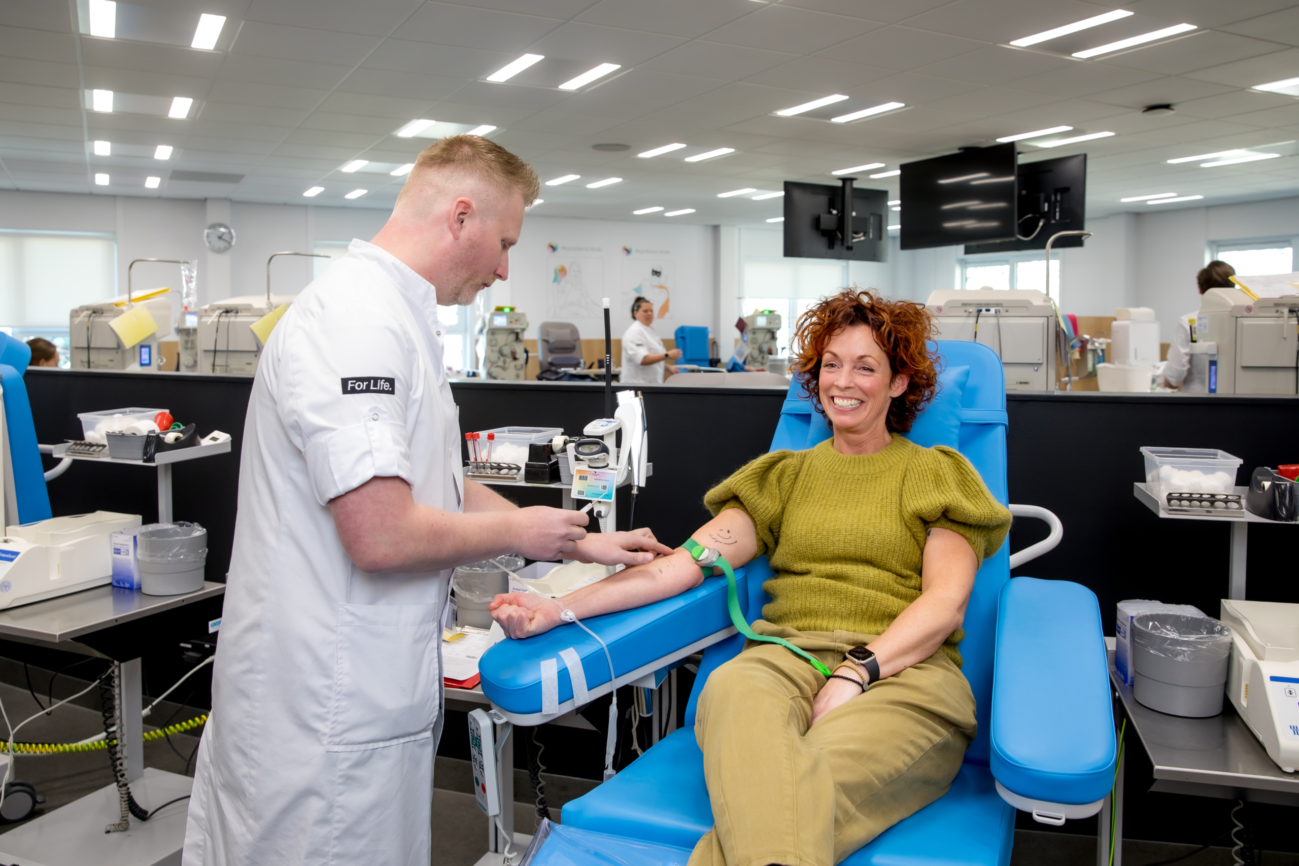 Een vrouw doneert bloed in een donatiecentrum, begeleid door een mannelijke medewerker in een witte jas.