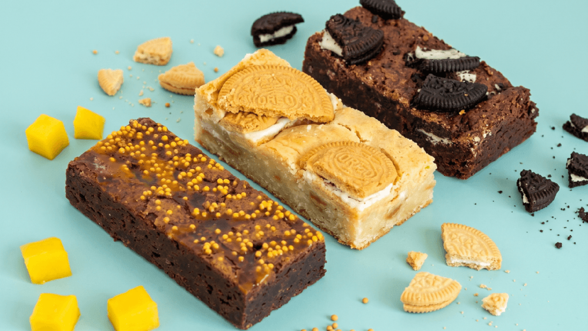 Drie verschillende brownies versierd met koekjes en chocolade liggen op een blauwe achtergrond, omringd door koekstukjes en gele blokjes.