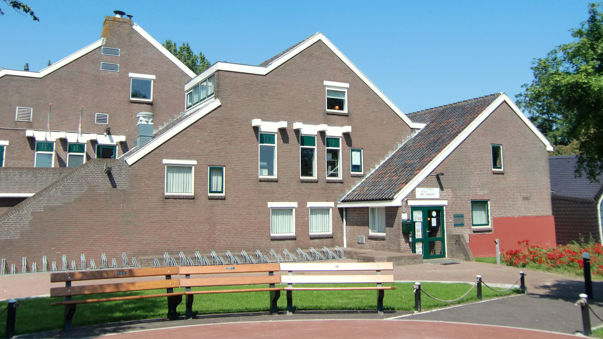 Bakstenen gebouw met een zadeldak en meerdere ramen, omgeven door groen en banken op de voorgrond.