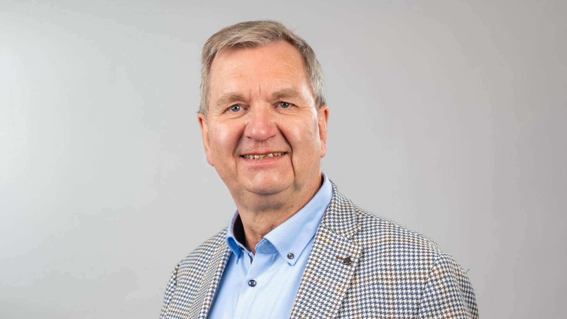 Man in een geruit jasje en lichtblauw overhemd kijkt lachend in de camera met een grijze achtergrond.