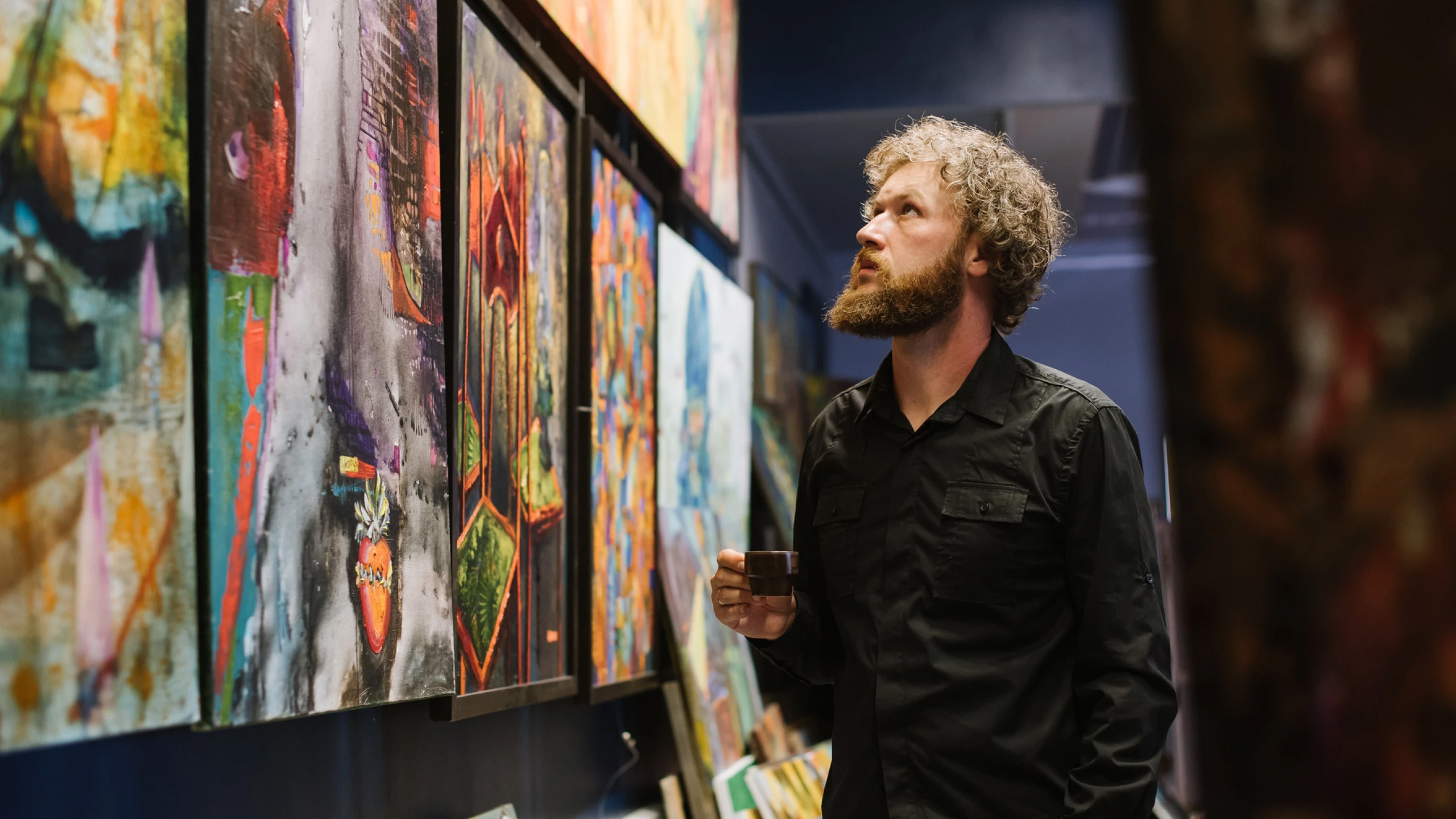 Een man met krullend haar en een baard bekijkt schilderijen in een kunstgalerie, terwijl hij een espressokopje vasthoudt.