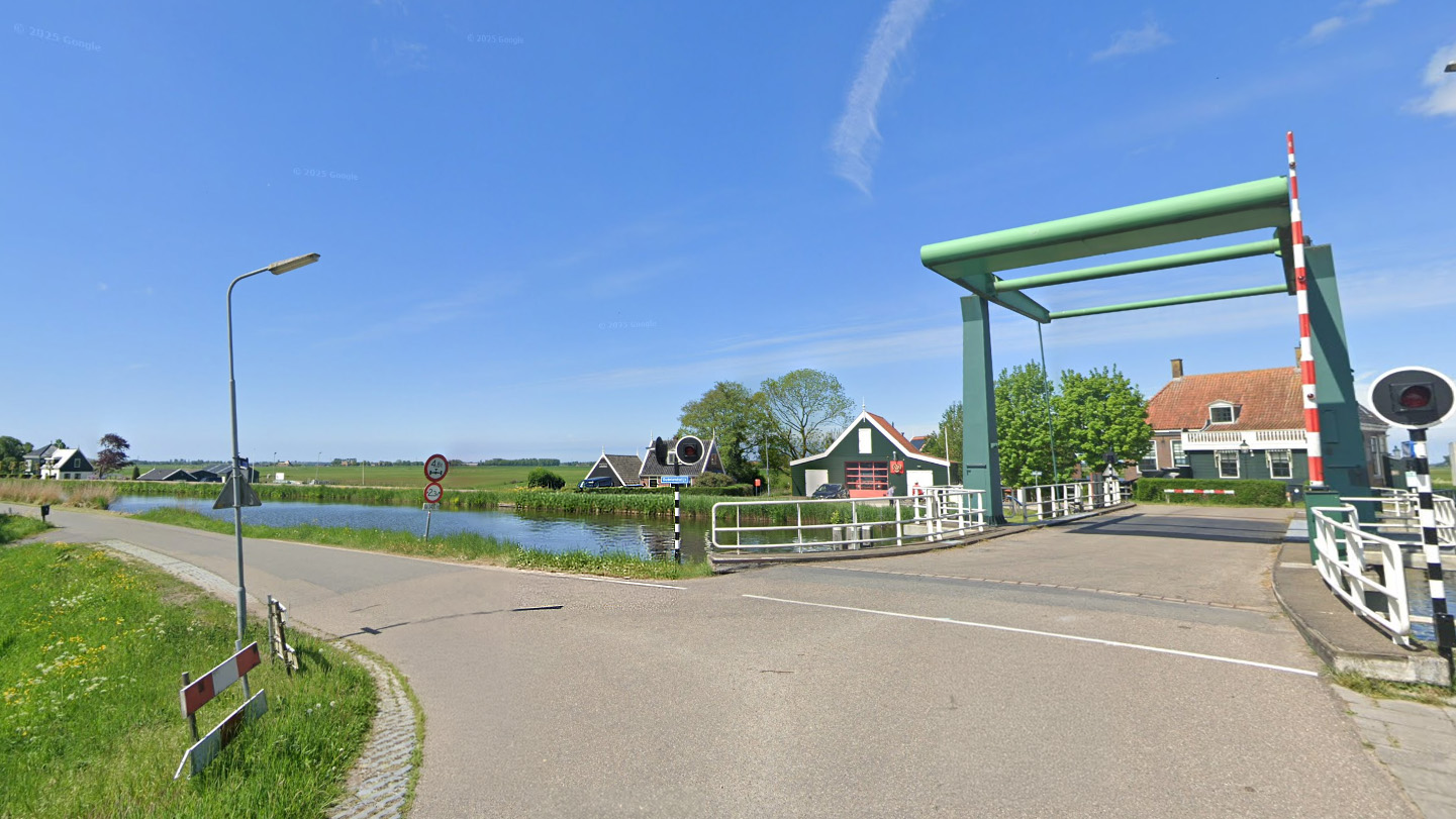 Groene ophaalbrug over een waterweg, omgeven door traditionele Nederlandse huizen en een weids landschap onder een blauwe lucht.