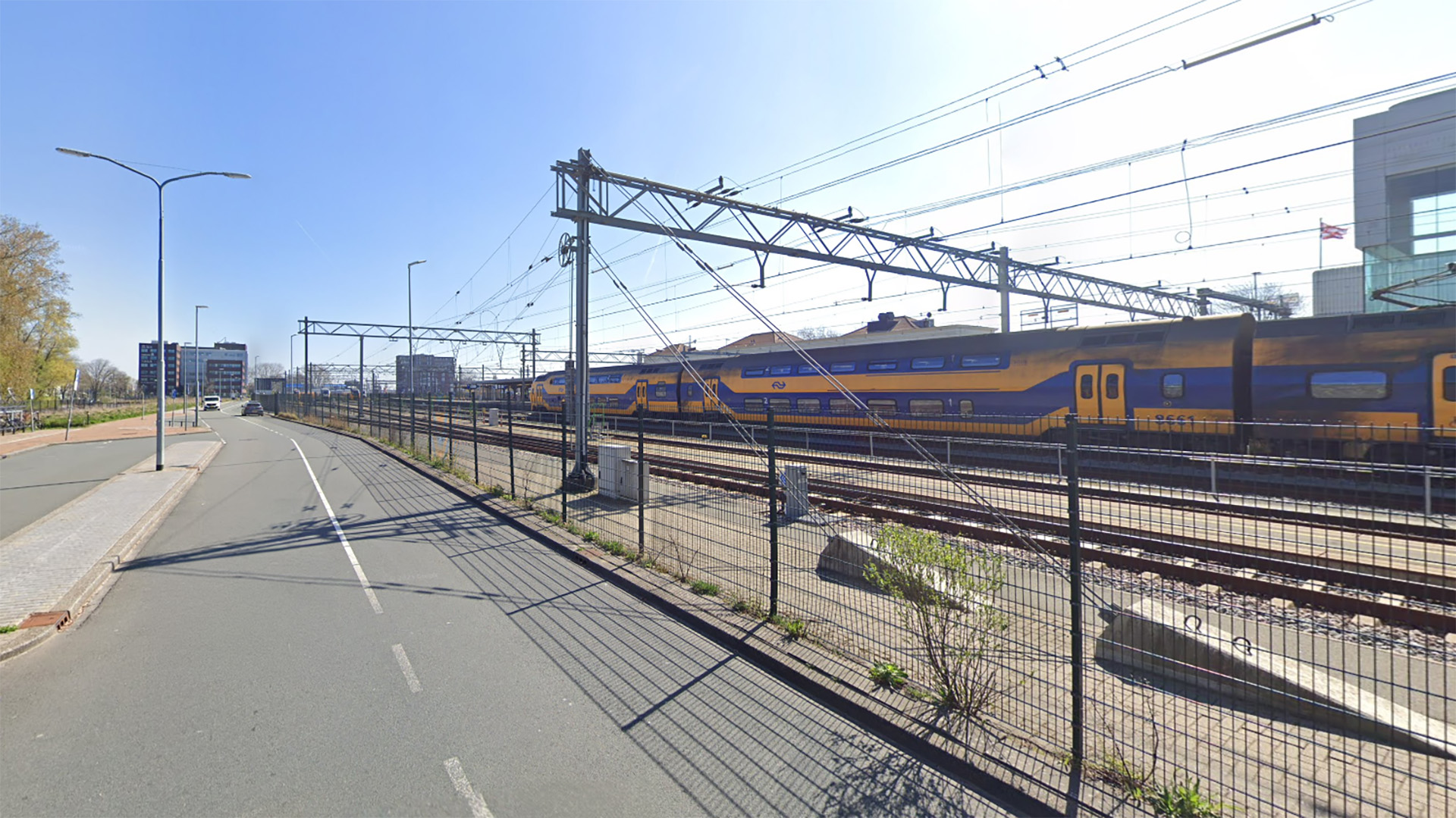 Een gele en blauwe dubbeldekstrein van de Nederlandse Spoorwegen langs een weg met fiets- en wandelpaden, onder een heldere lucht.