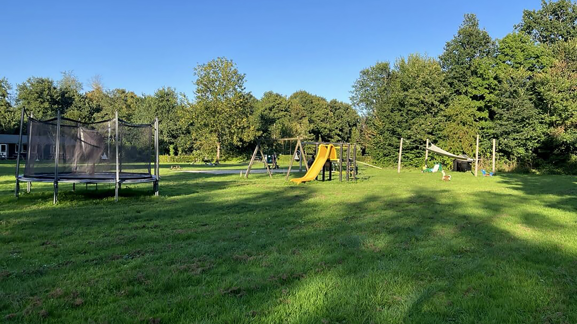 Speelplaats met een trampoline, glijbaan en open groene ruimte omringd door bomen onder een blauwe lucht.