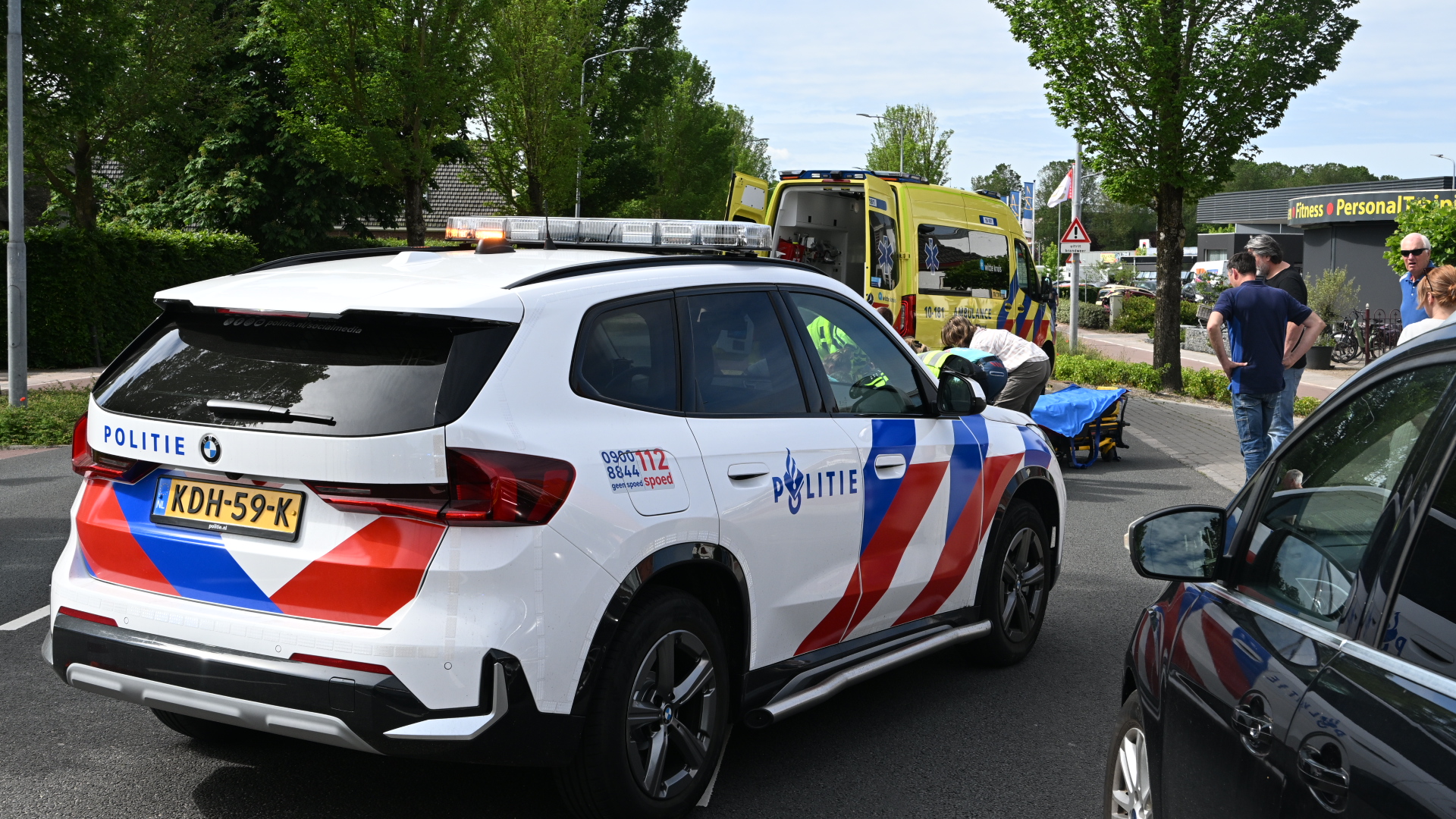 Politieauto en ambulance bij een incident, met hulpverleners en omstanders op straat.