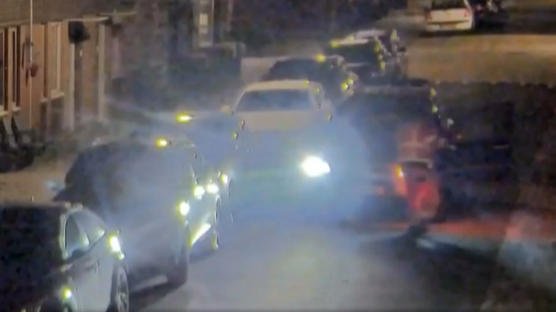 Auto's geparkeerd langs een donkere straat, met koplampen aan en lichte reflecties zichtbaar.