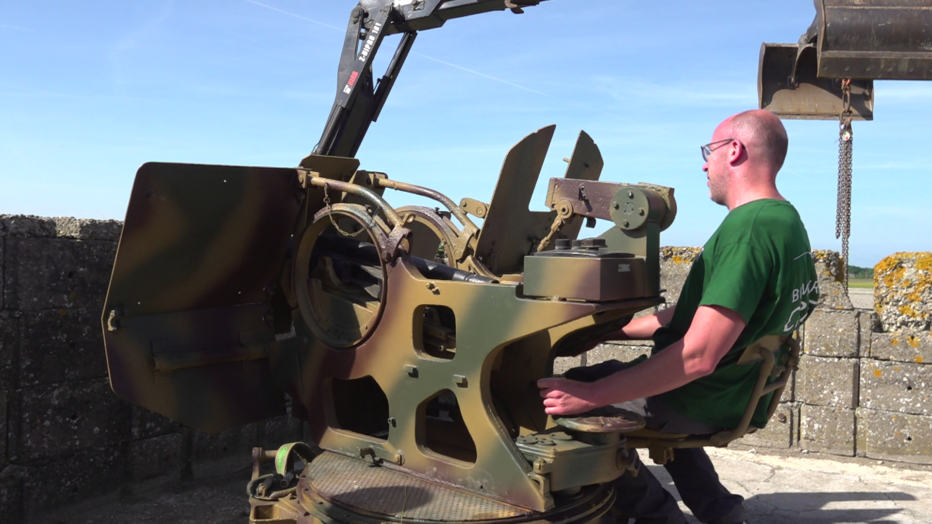 Man zittend op een oude militaire geschutsstelling, met een groene T-shirt en een heldere lucht op de achtergrond.