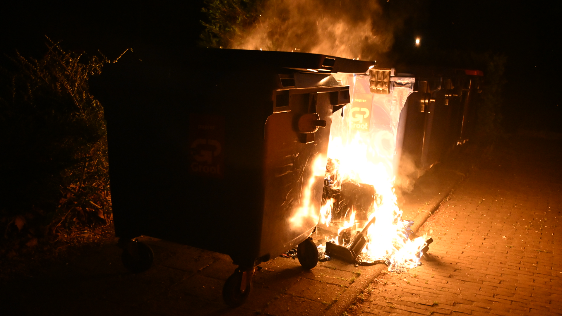 Brandende afvalcontainer naast een straat, met vlammen zichtbaar tegen de nachtelijke achtergrond.