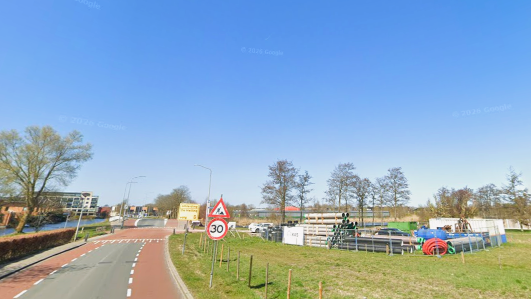 Weg met een snelheidsbord van 30 km/u, omringd door groen terrein en enkele bomen onder een heldere blauwe hemel.