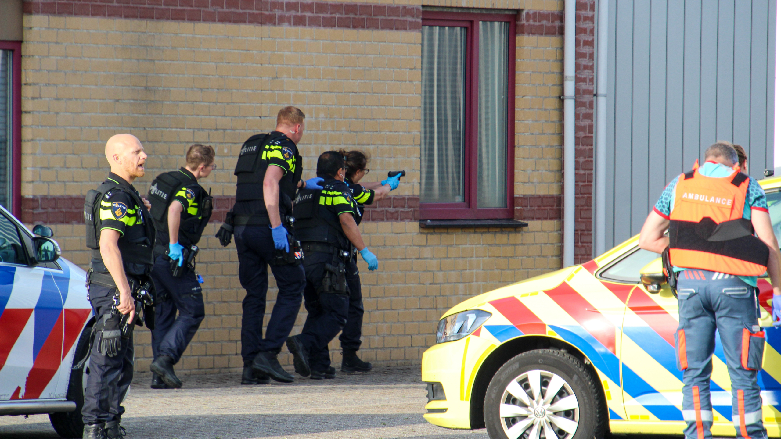 Politieagenten met handschoenen en apparatuur reageren bij een gebouw, naast een ambulancevoertuig.
