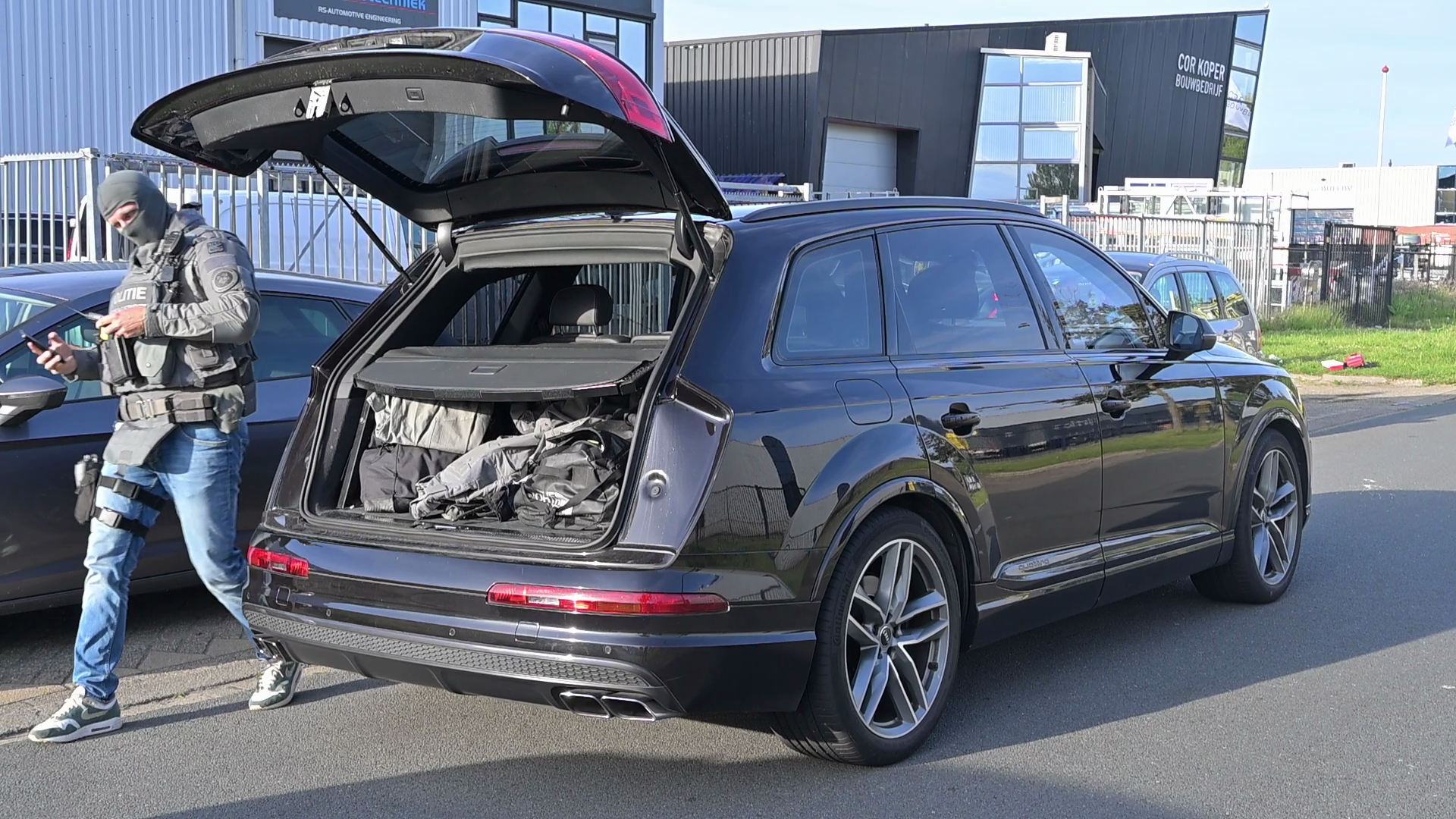 Een politieagent in tactische uitrusting staat naast een geopende kofferbak van een zwarte SUV op een industrieel terrein.
