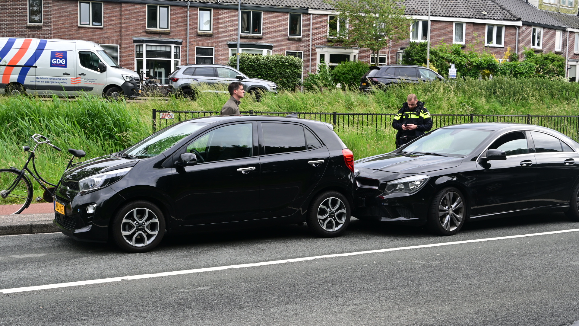 Twee zwarte auto's betrokken bij een kop-staartbotsing naast een fietspad; een politieagent kijkt op zijn telefoon.