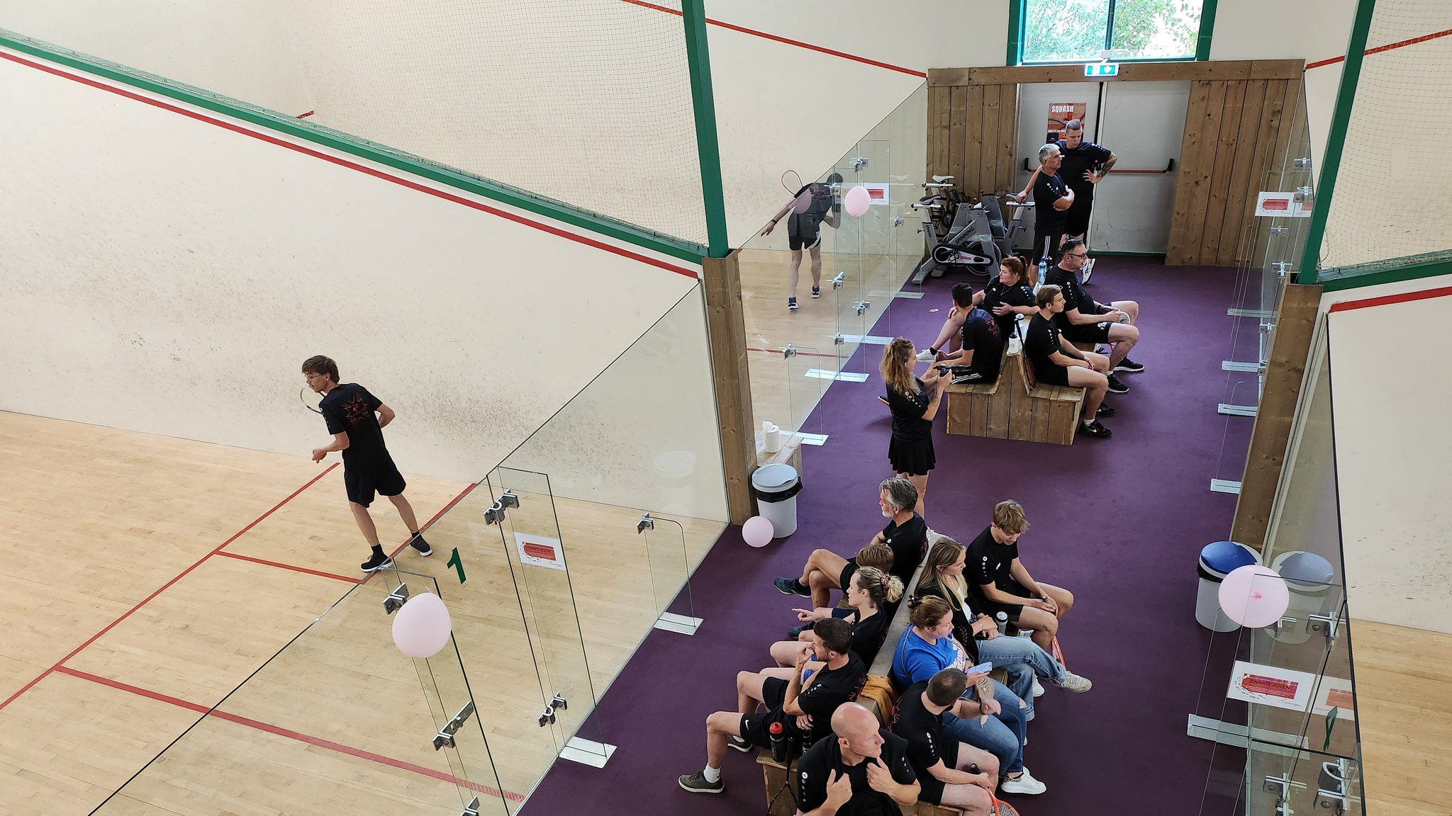 Mensen kijken toe bij een squashwedstrijd; een speler serveert op de baan met een glazen omheining.