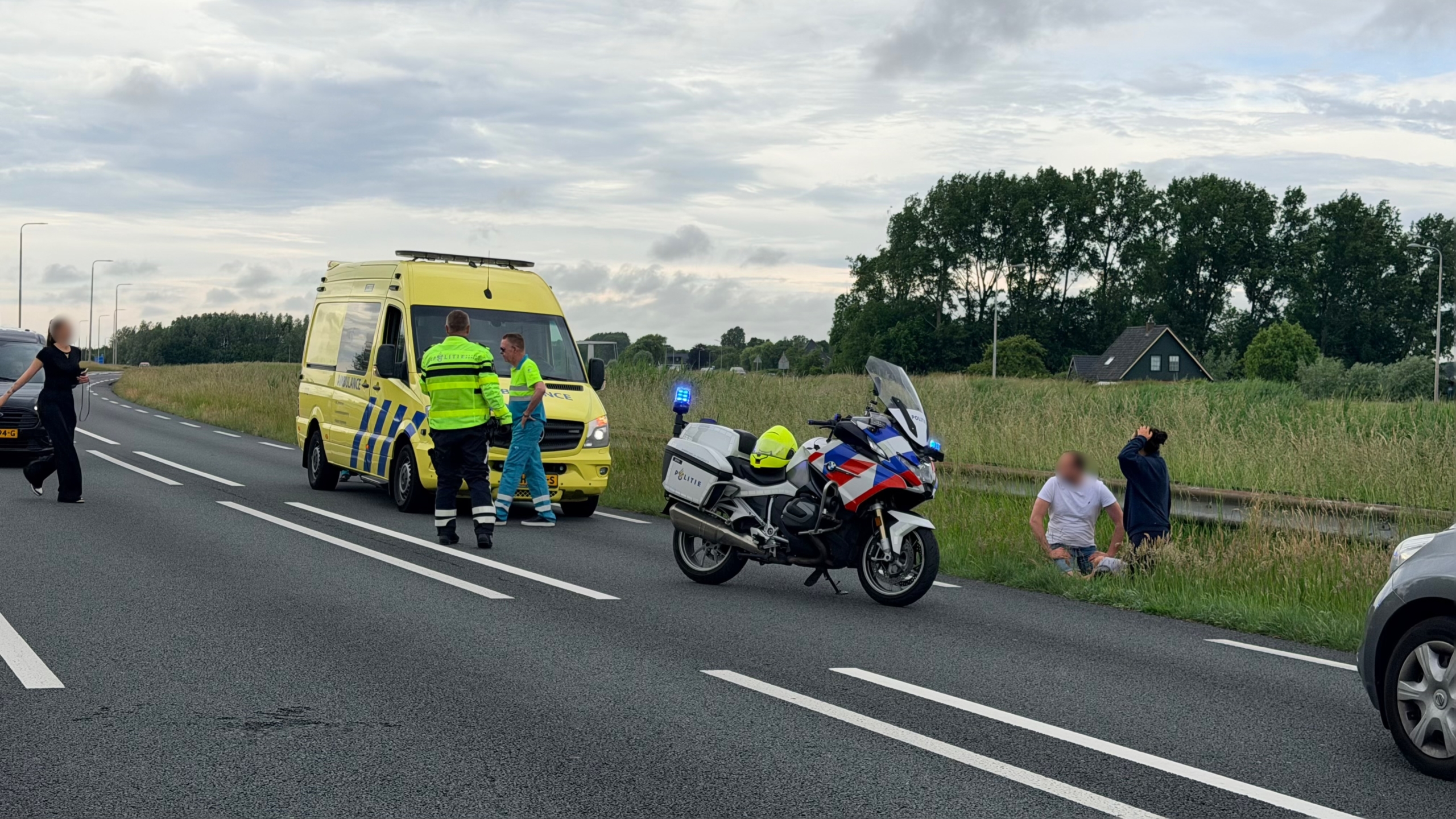 Ambulance en politiemotor op snelweg, met twee personen zittend langs de berm, terwijl hulpverleners overleggen.