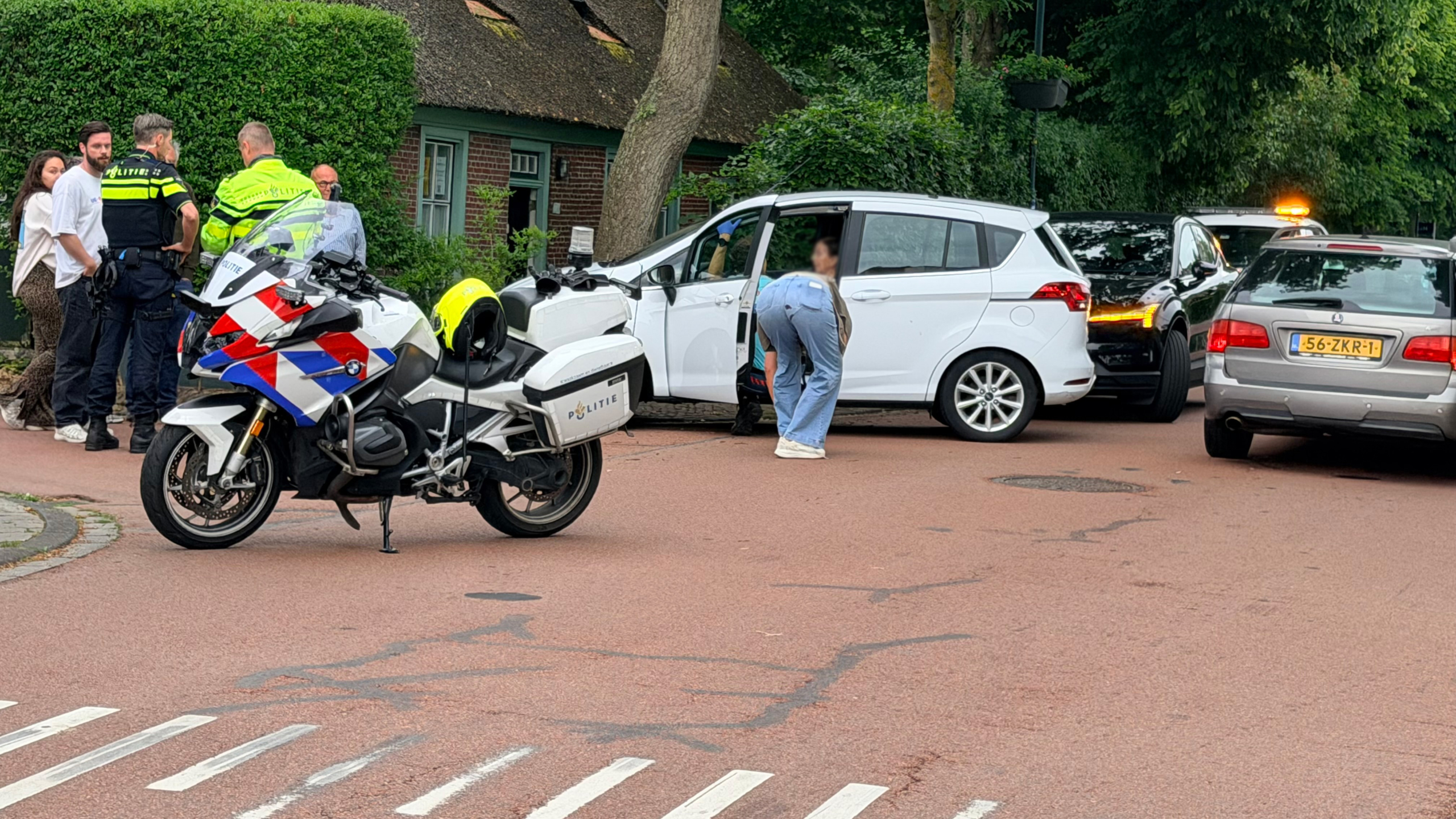 Politieagenten overleggen op straat bij een wit voertuig, met een politie-motor en meerdere auto's op de achtergrond.