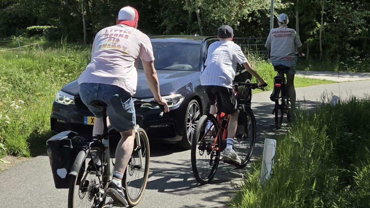 Drie mensen fietsen op een smal pad terwijl er een auto wacht.