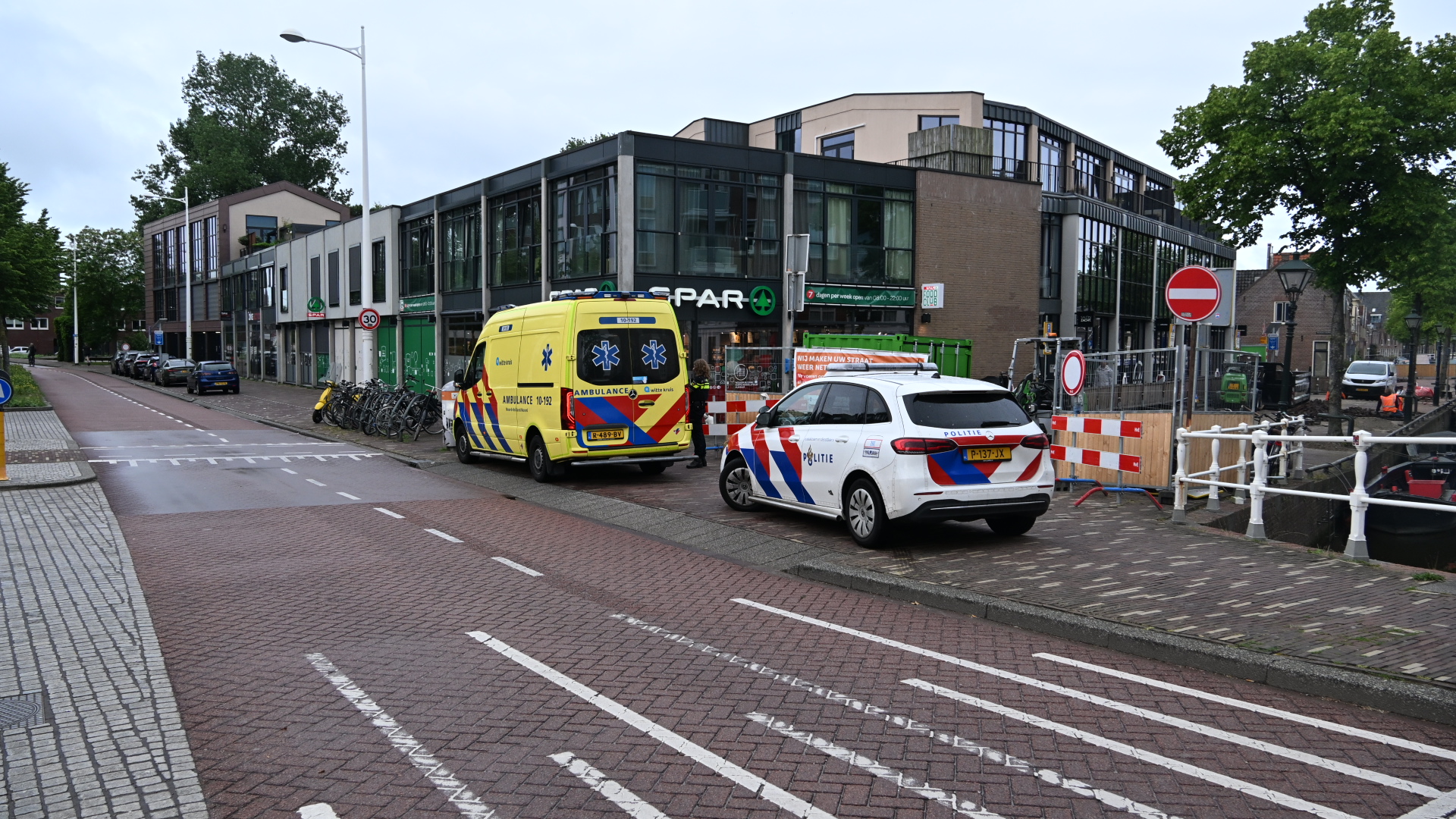 Een ambulance en politieauto staan geparkeerd bij een supermarkt in een straat met wegafzettingen en verbodsborden.