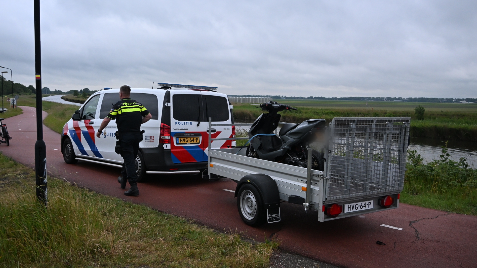 Een politieagent bij een politieauto met aanhanger waarop een scooter staat, langs een fietspad in een landelijke omgeving.