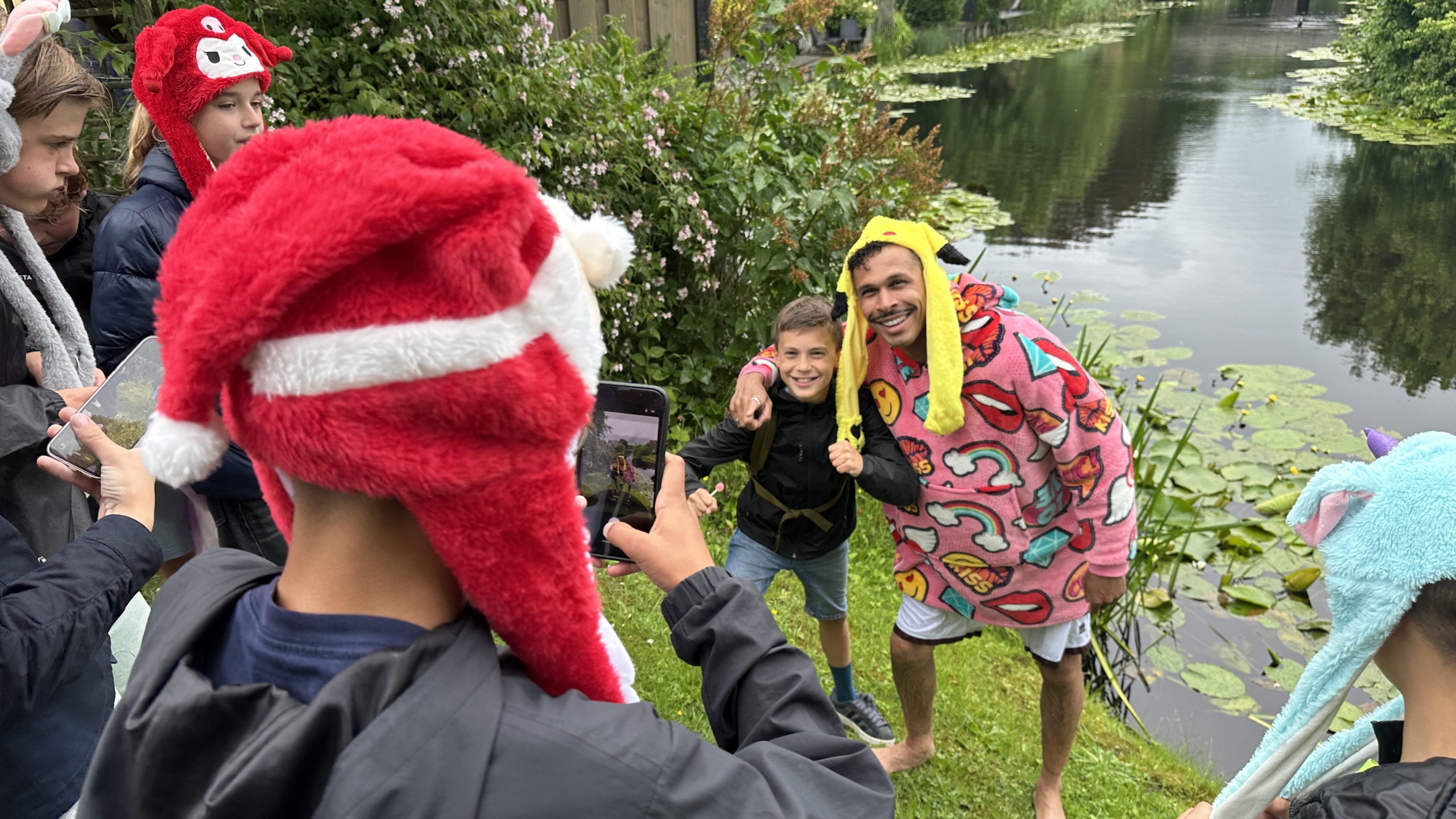 Een lachende man en een jongen poseren voor een foto aan de oever van een vijver, omringd door mensen in vrolijke dierenmutsen.