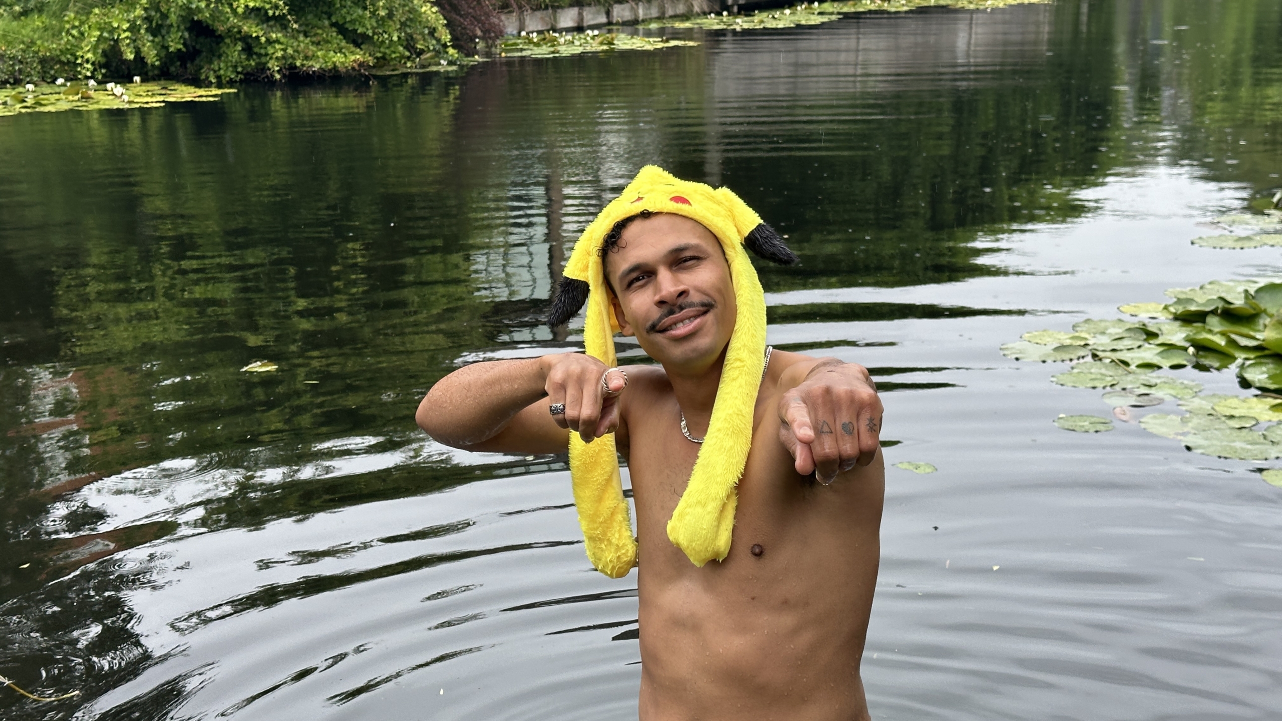 Persoon in een vijver draagt een gele Pikachu-hoed en wijst glimlachend naar de camera, omringd door waterplanten.