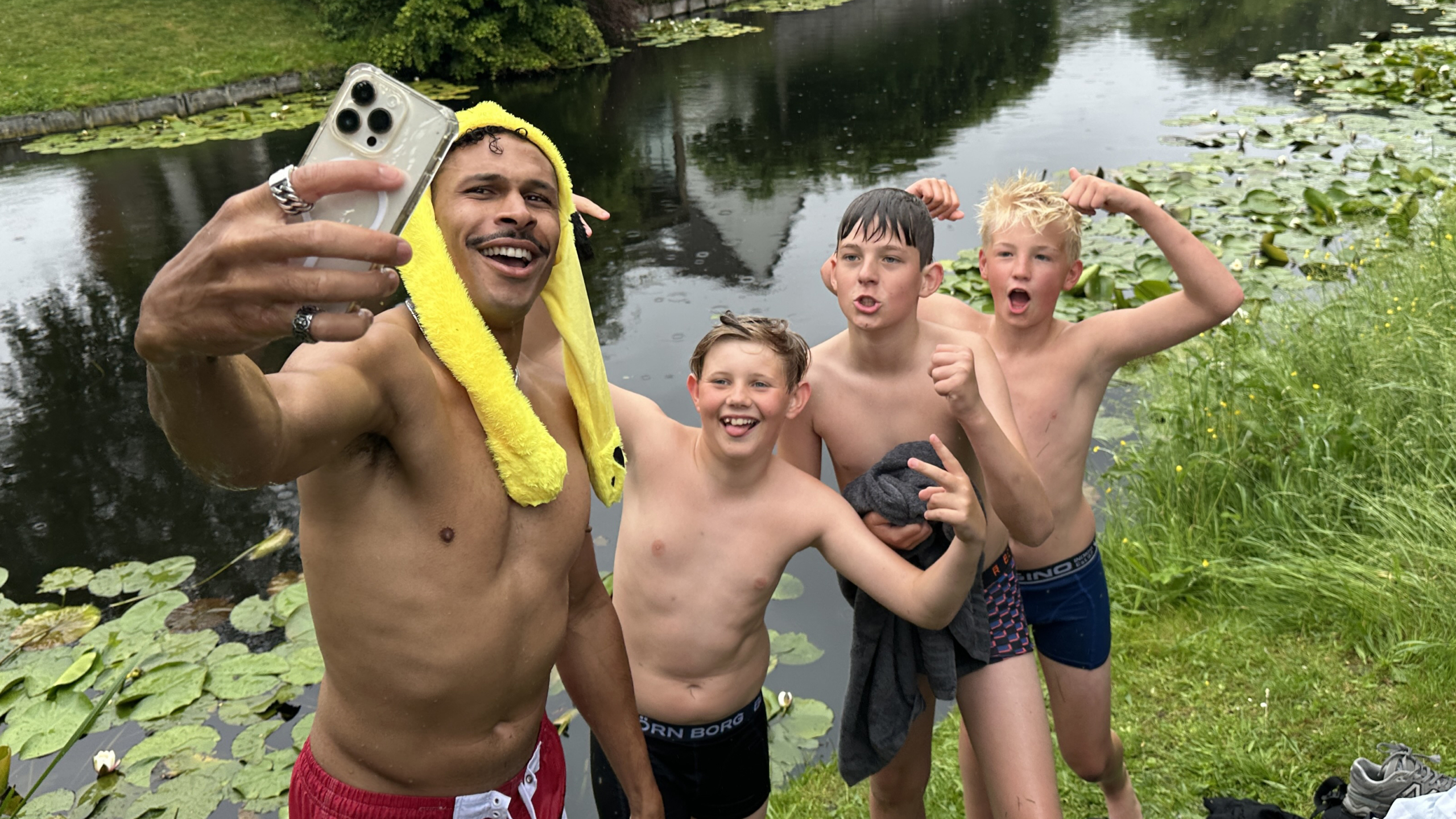 Een man met een gele handdoek op zijn hoofd maakt een selfie met drie lachende jongens bij de waterkant.