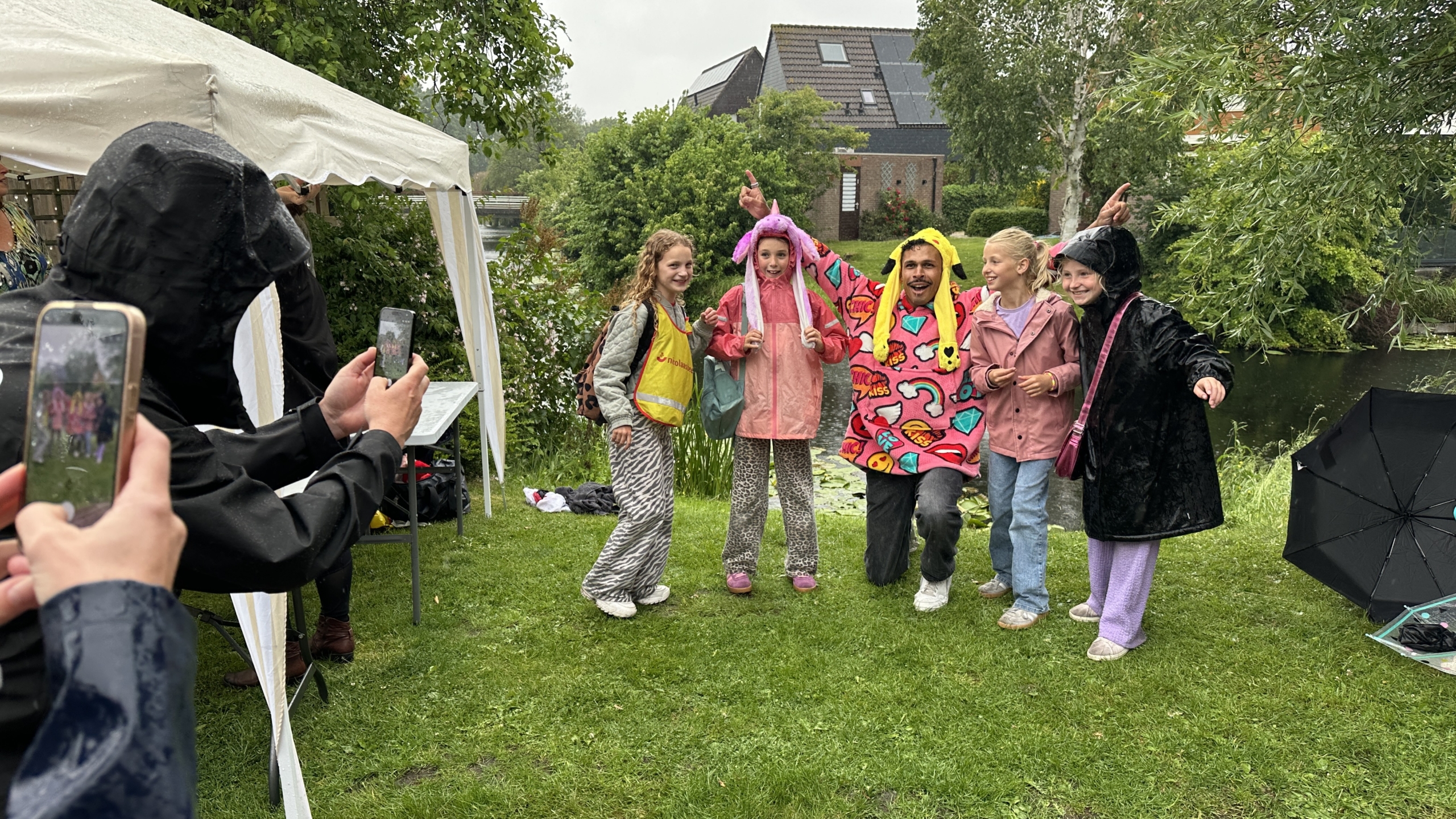 Groep kinderen en een volwassene poseren in kleurrijke regenkleding terwijl een foto wordt gemaakt, bij een tent in een regenachtig park.