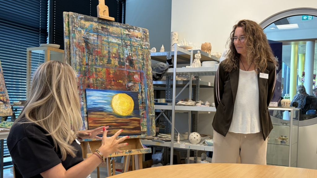 Een vrouw zit voor een schildersezel en bespreekt een schilderij van een zon met een staande vrouw in een atelier.