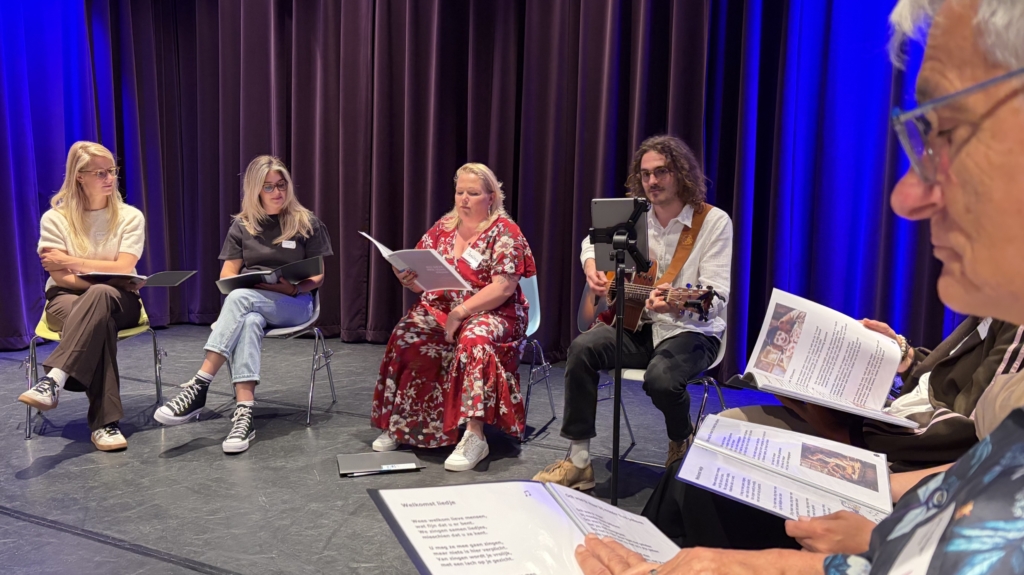 Een groep mensen zit in een kring, sommigen lezen muziekbladen, terwijl een man gitaar speelt op een podium met blauwe verlichting.
