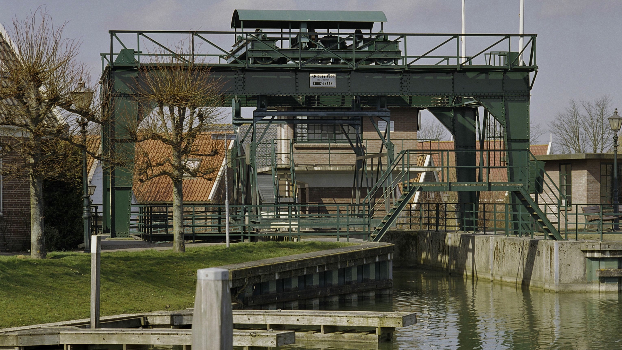 Historische metalen ophaalbrug over een waterkanaal met omringende woningen en kale bomen.