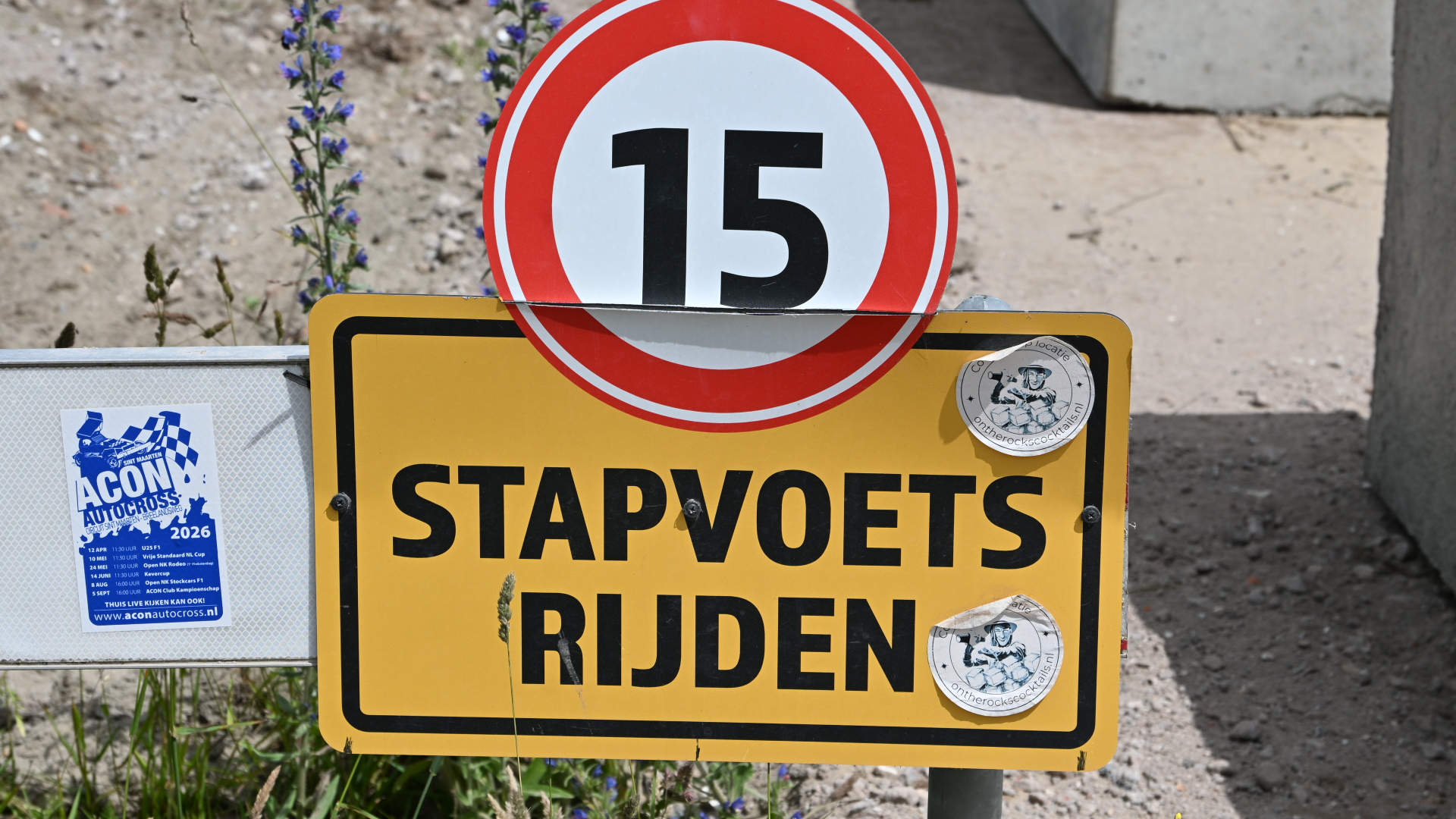 Verkeersbord met snelheidslimiet van 15 km/u en de tekst "Stapvoets rijden", met stickers en een poster op de achtergrond.