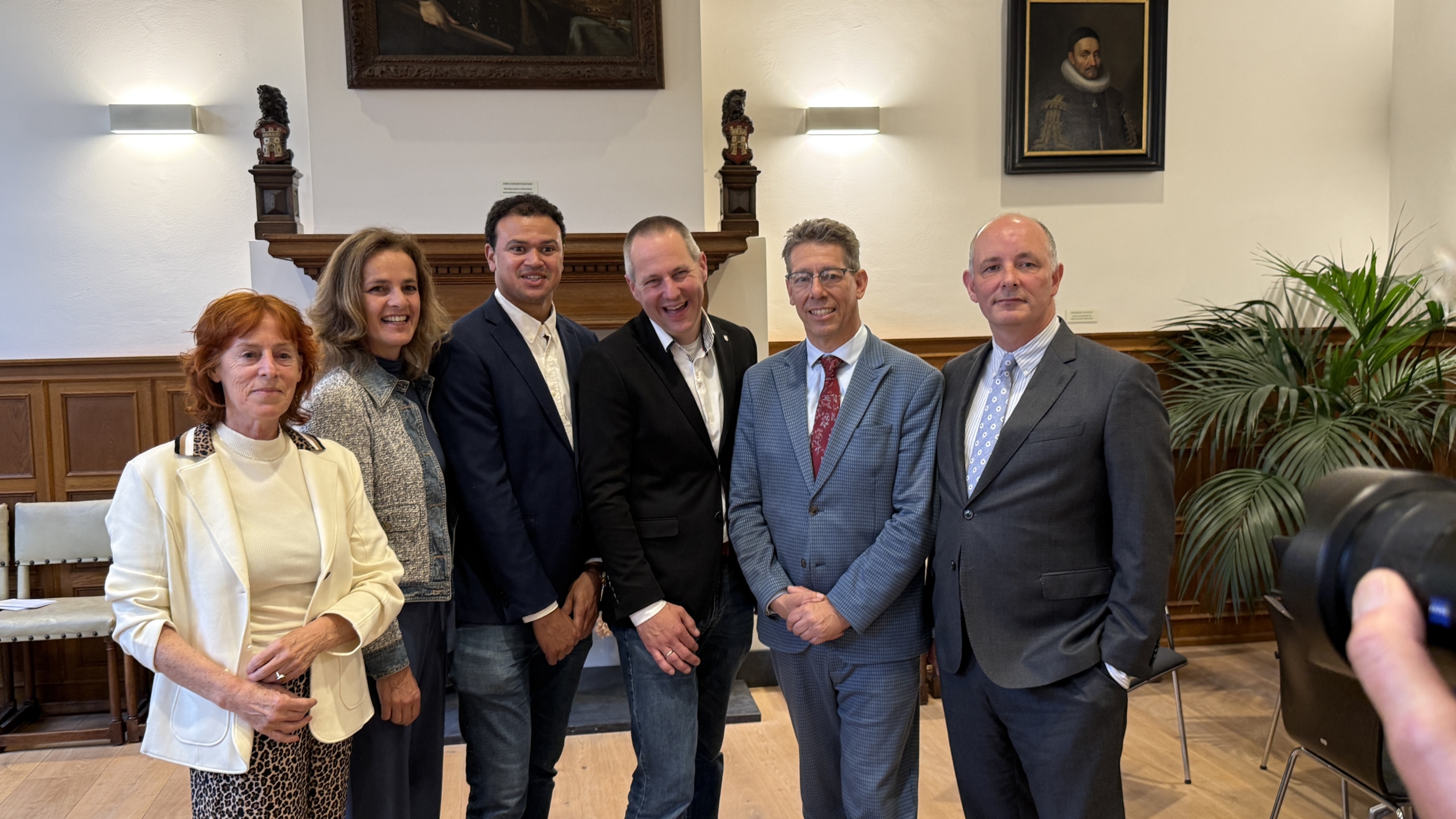 Zes mensen poseren samen in een klassiek ingerichte kamer met houten lambrisering en schilderijen aan de muur.