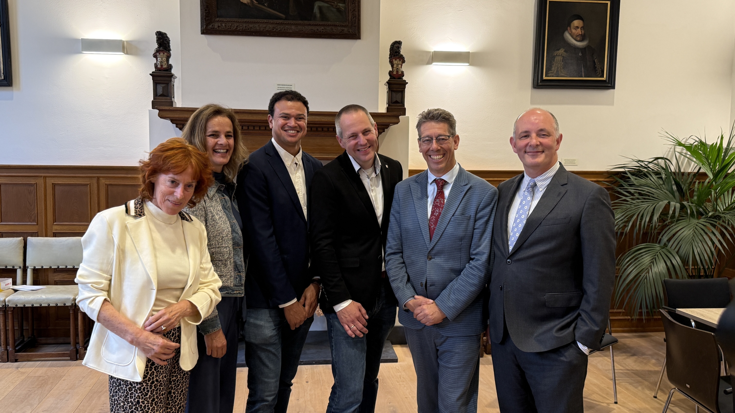 Zes mensen poseren lachend in een stijlvolle kamer met houten panelen en portretten aan de muur.