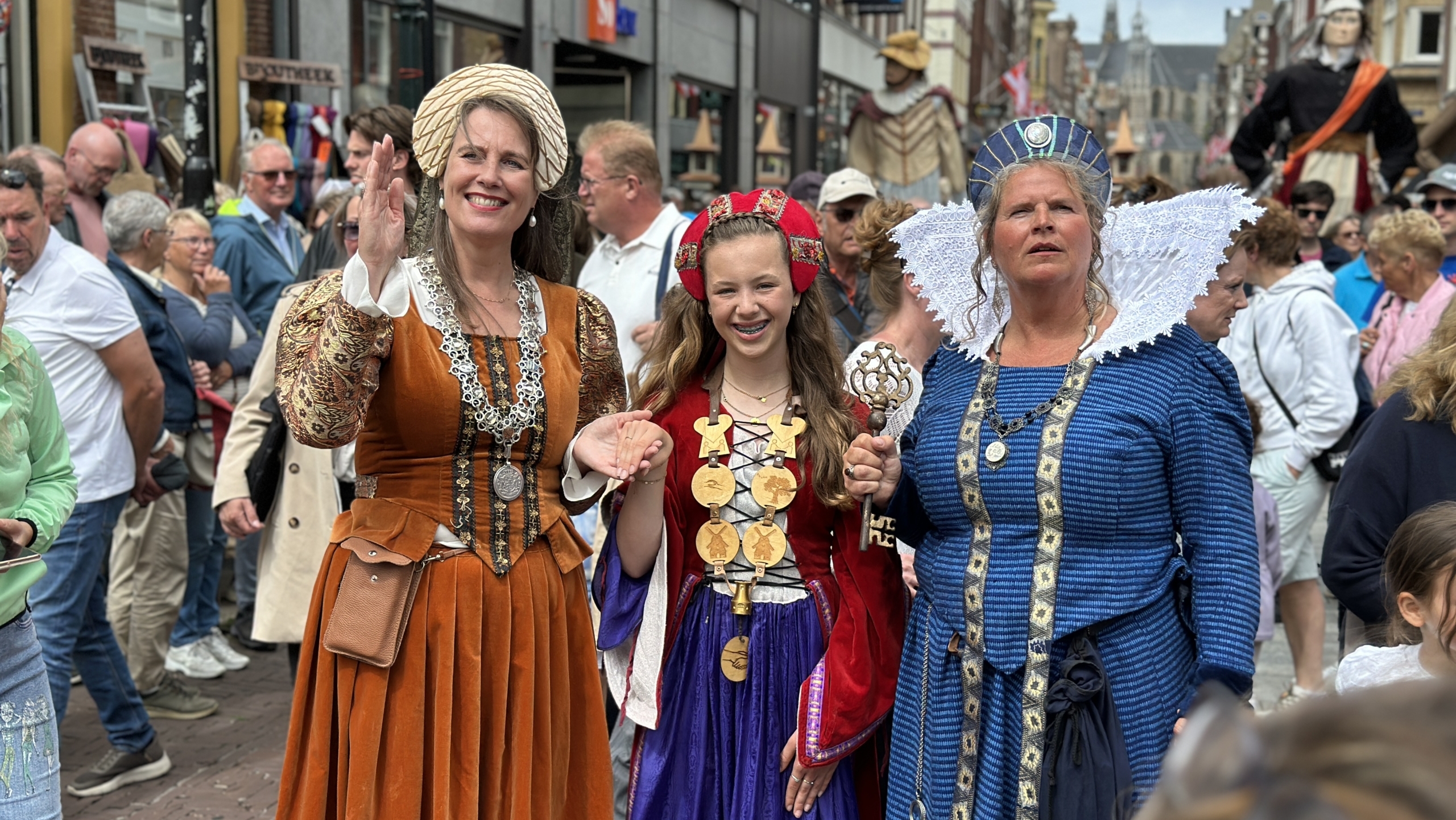 Drie vrouwen in historische kostuums poseren lachend tussen een menigte op een straatfestival.