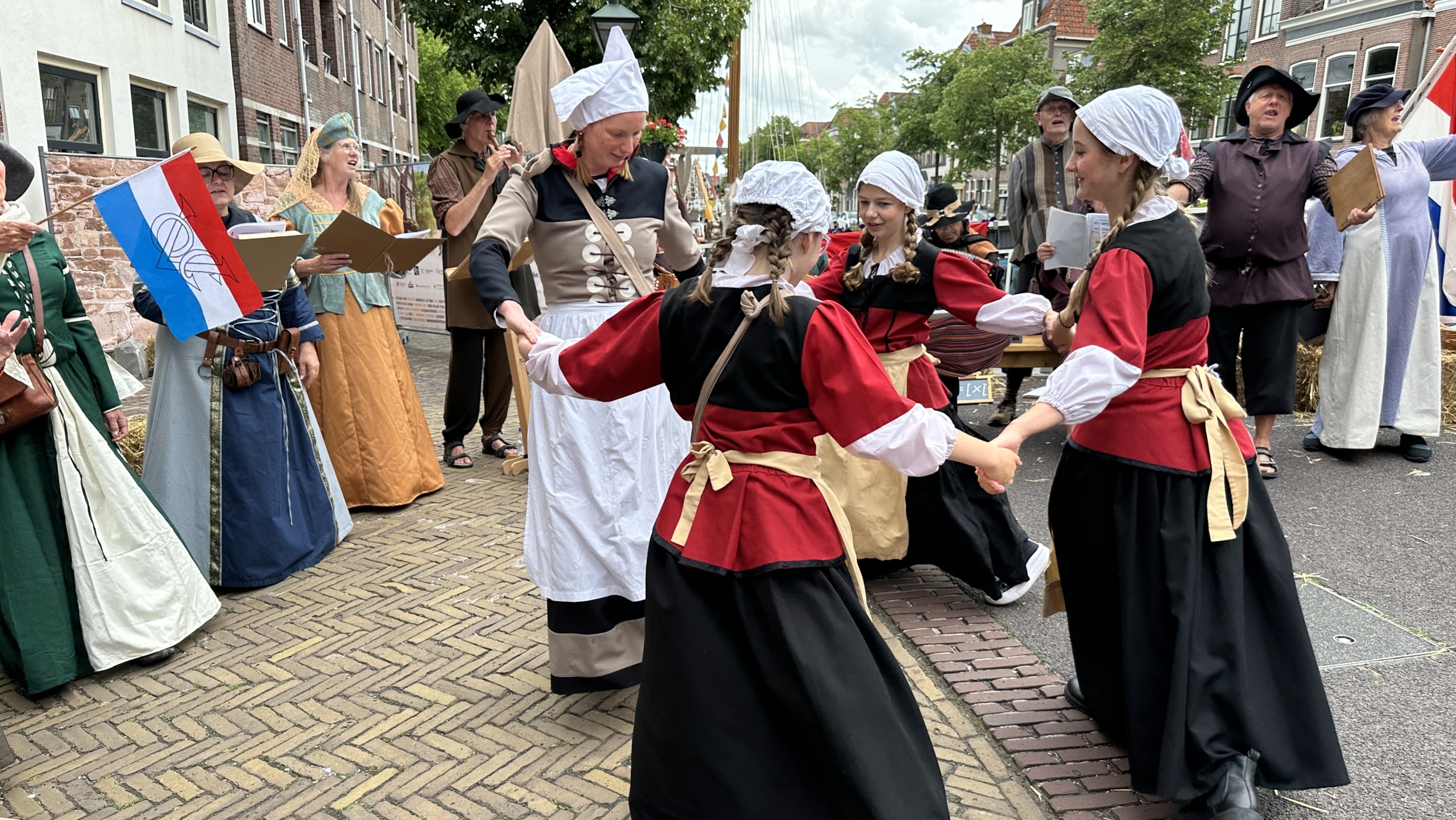 Mensen in traditionele kleding dansen en zingen op een straatfestival, met Nederlandse vlaggen in de achtergrond.