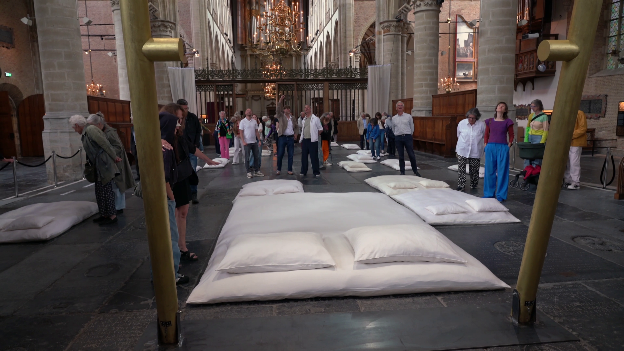 Groep mensen in een kerkachtige ruimte, kijkend naar grote witte kussens en matrassen op de vloer, tussen pilaren en kroonluchters.