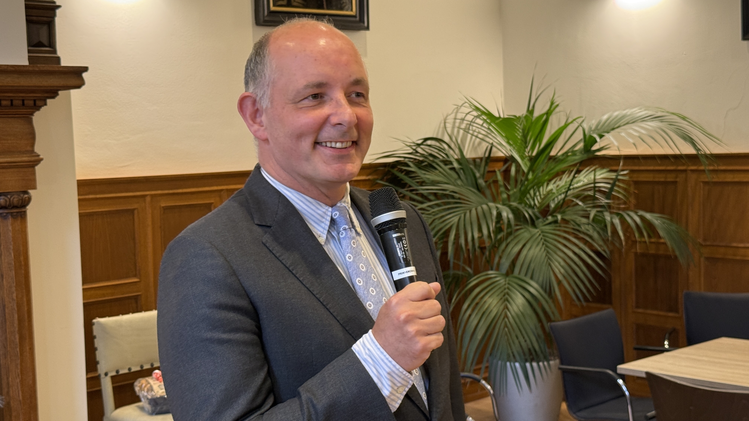 Man in een pak houdt een microfoon vast in een zaal met houten lambrisering en een groene plant op de achtergrond.