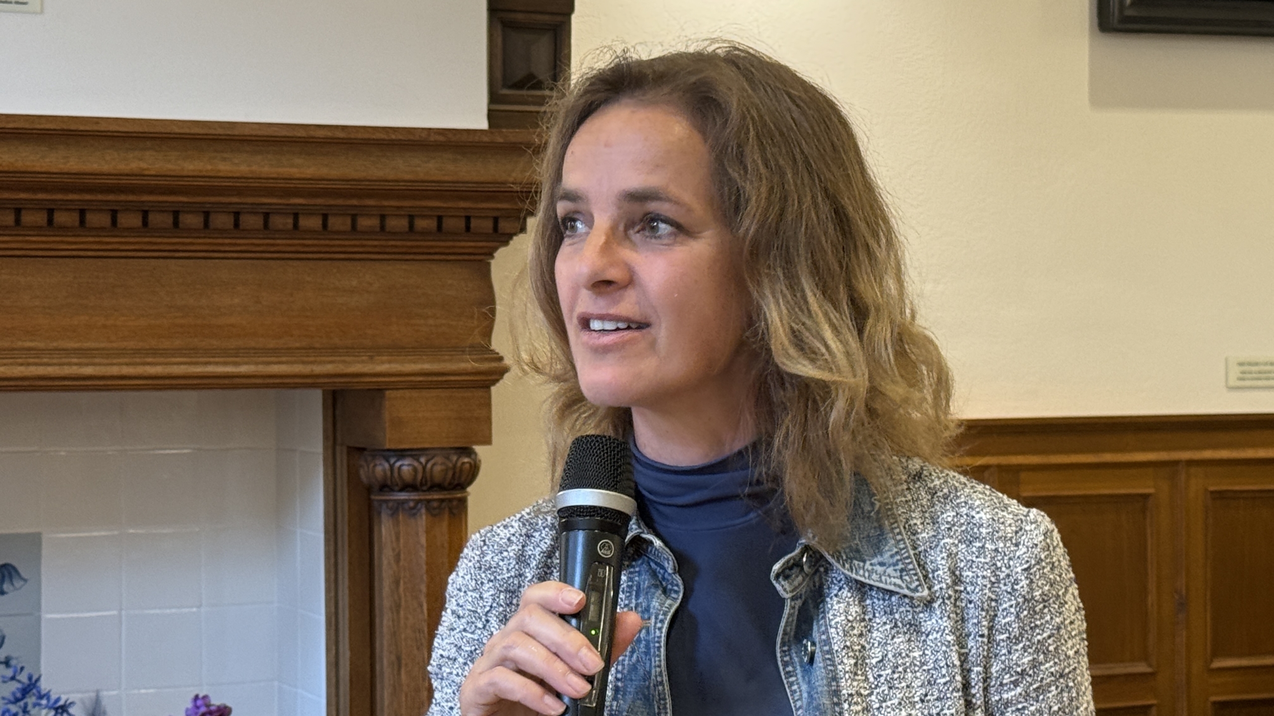 Vrouw met halflang haar spreekt in een microfoon, in een kamer met houten lambrisering.