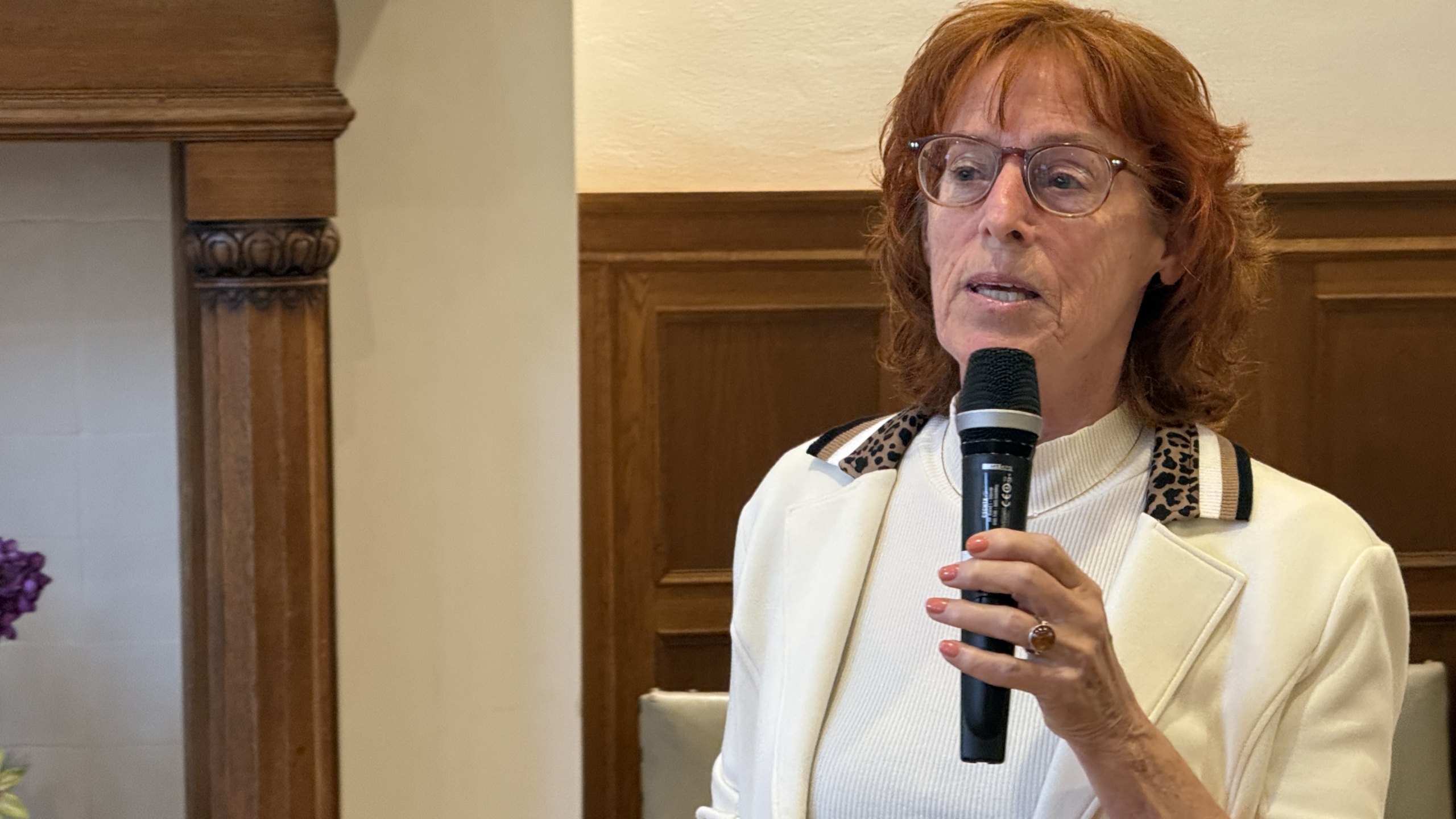 Vrouw met rood haar spreekt in een microfoon, gekleed in een witte jas met luipaardprint accenten, in een kamer met houten lambrisering.